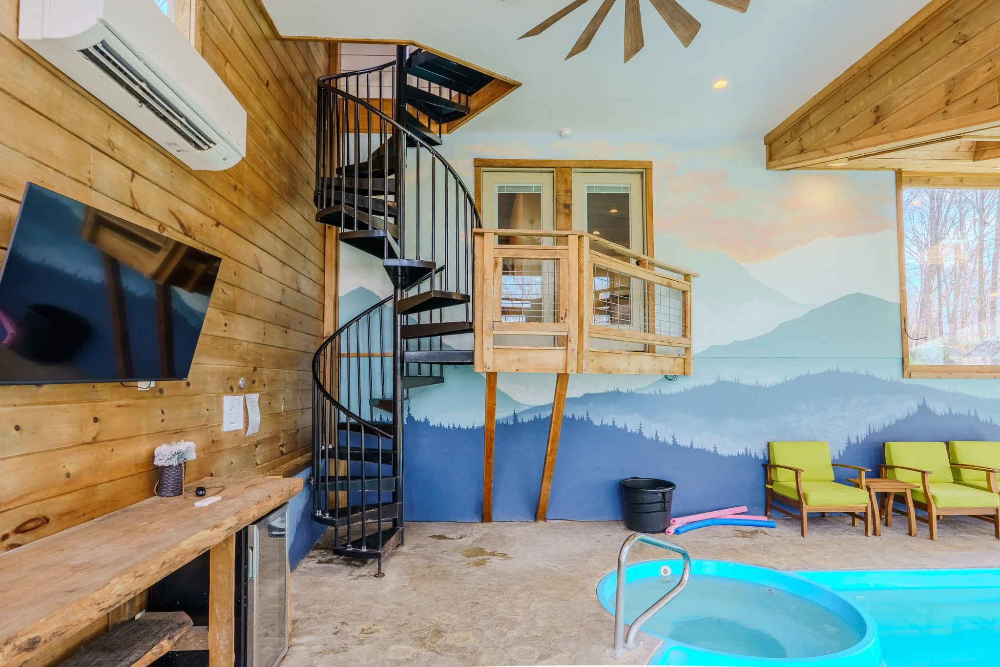 Indoor swimming pool area with a spiral staircase, a wall painted with mountain scenery, green chairs, a wooden balcony, and a large window showing trees outside.