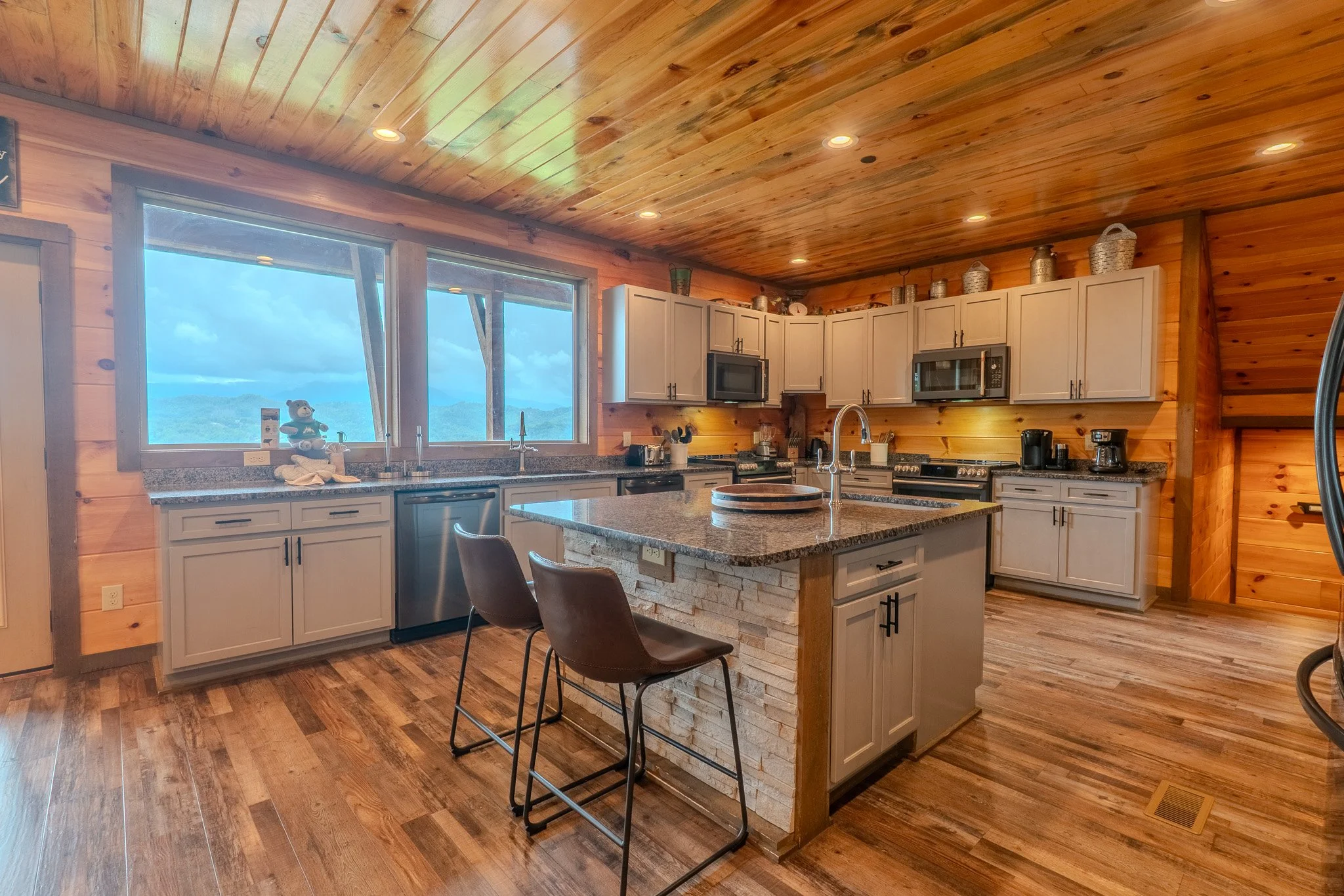 Kitchen with wooden walls and ceiling, granite countertops, island with stone base, white cabinets, stainless steel appliances, three brown chairs at the island, large windows with a view of mountains, and hardwood flooring.