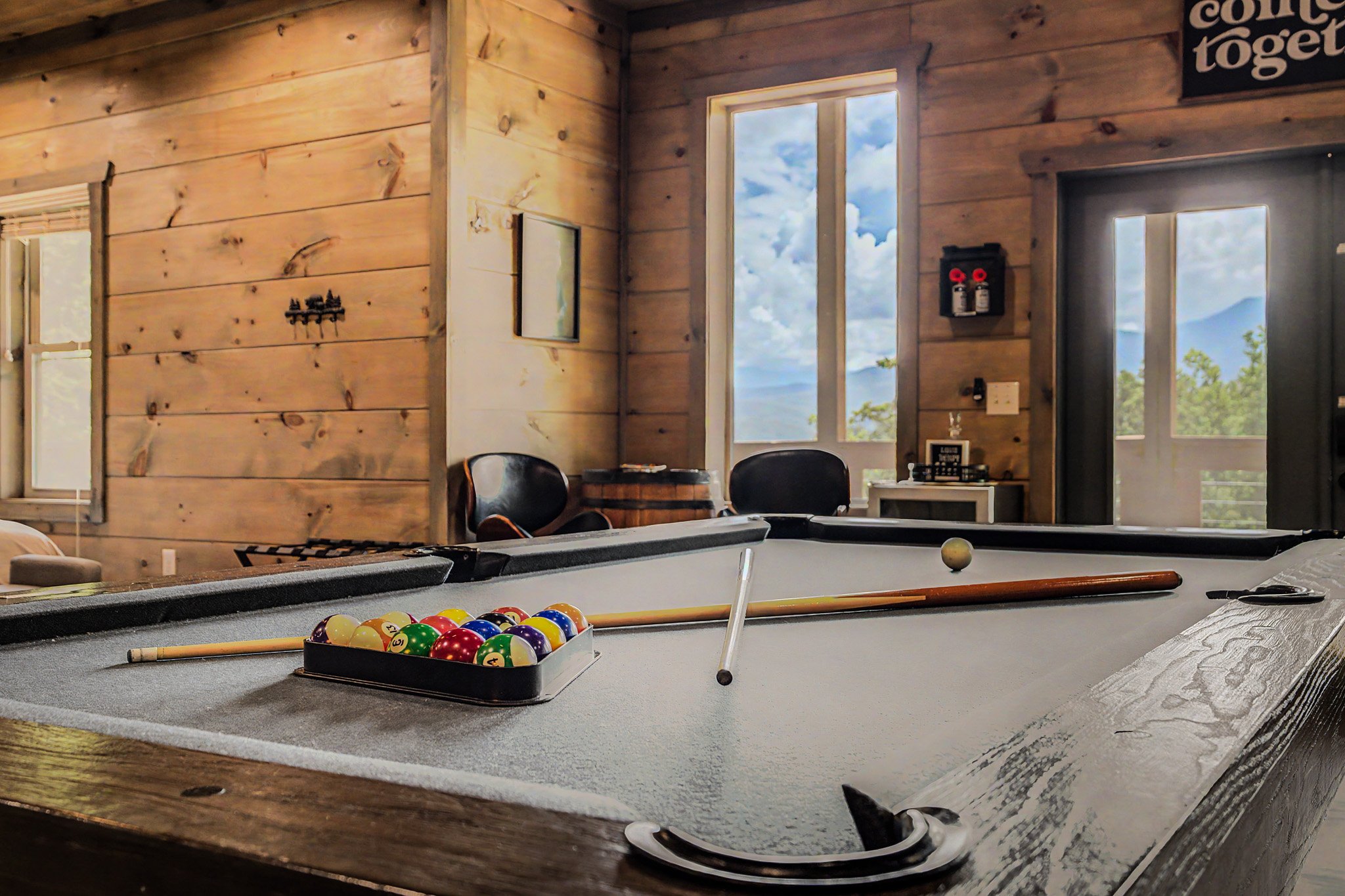 A pool table with a rack of billiard balls, a cue stick, and part of a white cue ball visible. The room has wooden paneled walls, large windows showing a sky with clouds and mountains outside, and some wall decorations including a framed picture and 