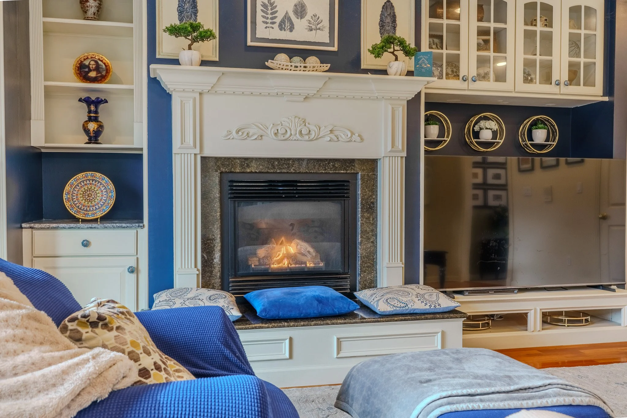 Living room with a fireplace surrounded by blue walls, decorated with plants, artwork, and various decorative items, with blue and patterned pillows on a sofa and a television on the right.