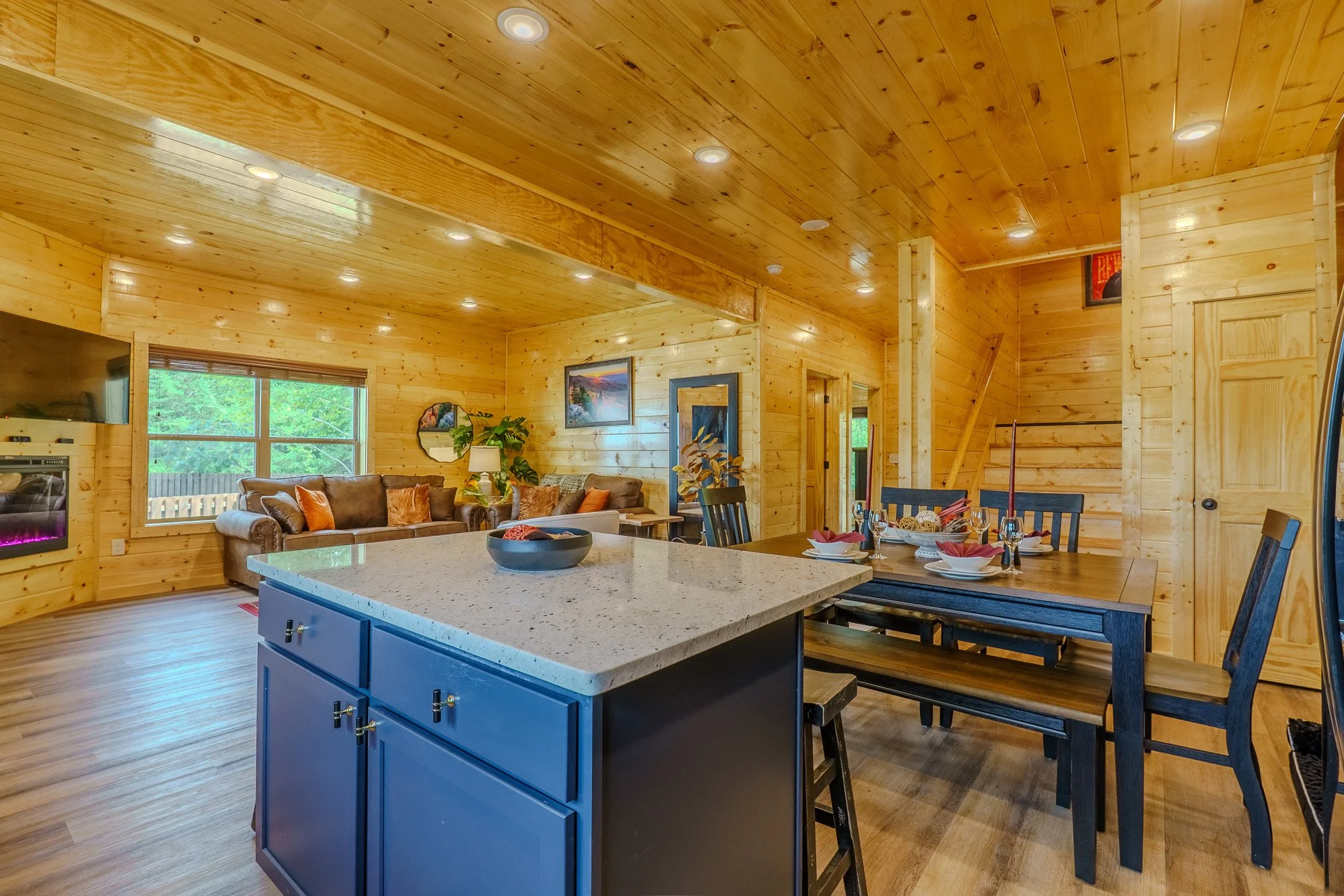 Open-concept living and dining area with wooden walls and ceiling, a kitchen island with a light-colored countertop, a dining table with black chairs and place settings, a sofa, window with green trees outside, wall art, and a staircase in the backgr