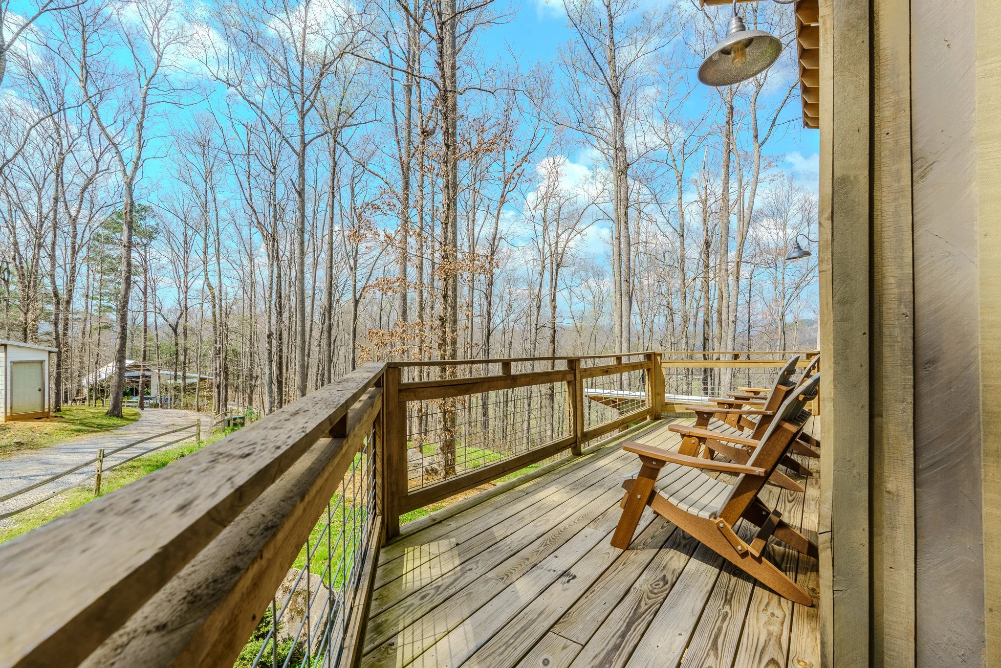 A wooden deck with four chairs faces a wooded area with tall, leafless trees and a partly cloudy blue sky.