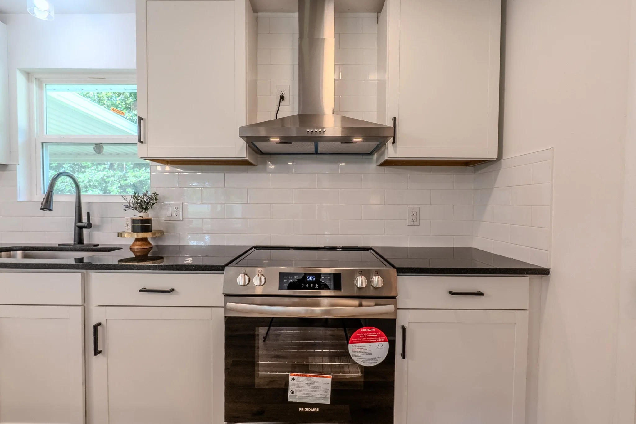 Modern kitchen with white cabinets, black countertops, a stainless steel oven with a glass door, a black range hood, white tile backsplash, a window with greenery outside, and a small potted plant on the counter.