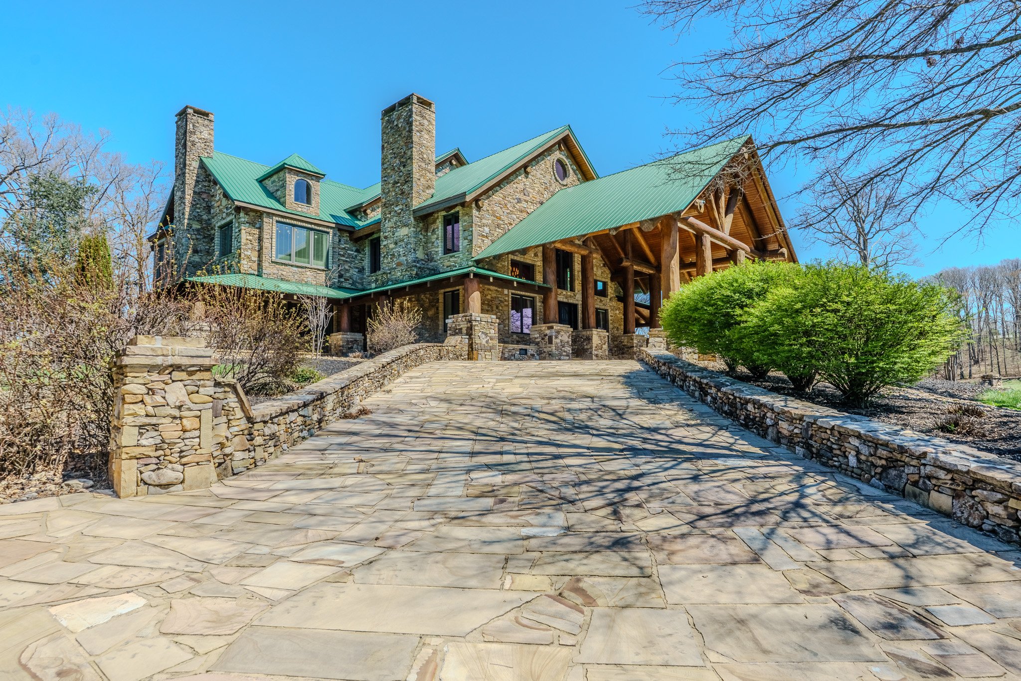 A large stone house with a green metal roof, multiple chimneys, and wooden accents, surrounded by landscaped bushes and trees, with a stone driveway leading up to it on a sunny day.