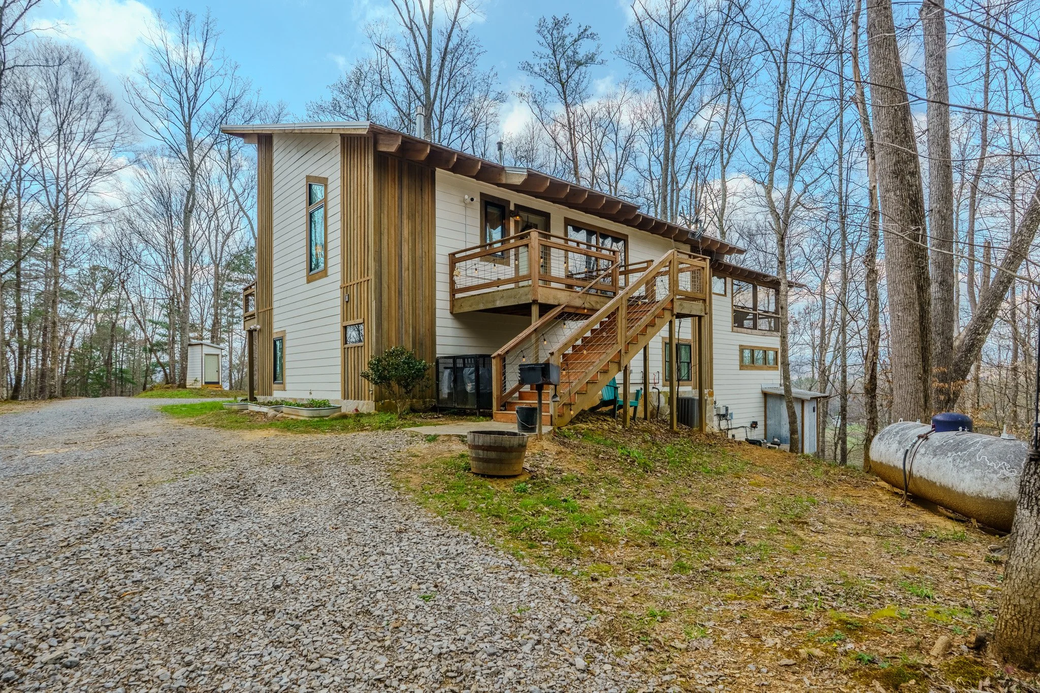 A modern two-story house with a wood and white exterior, a gravel driveway, and a wooded backyard with leafless trees.