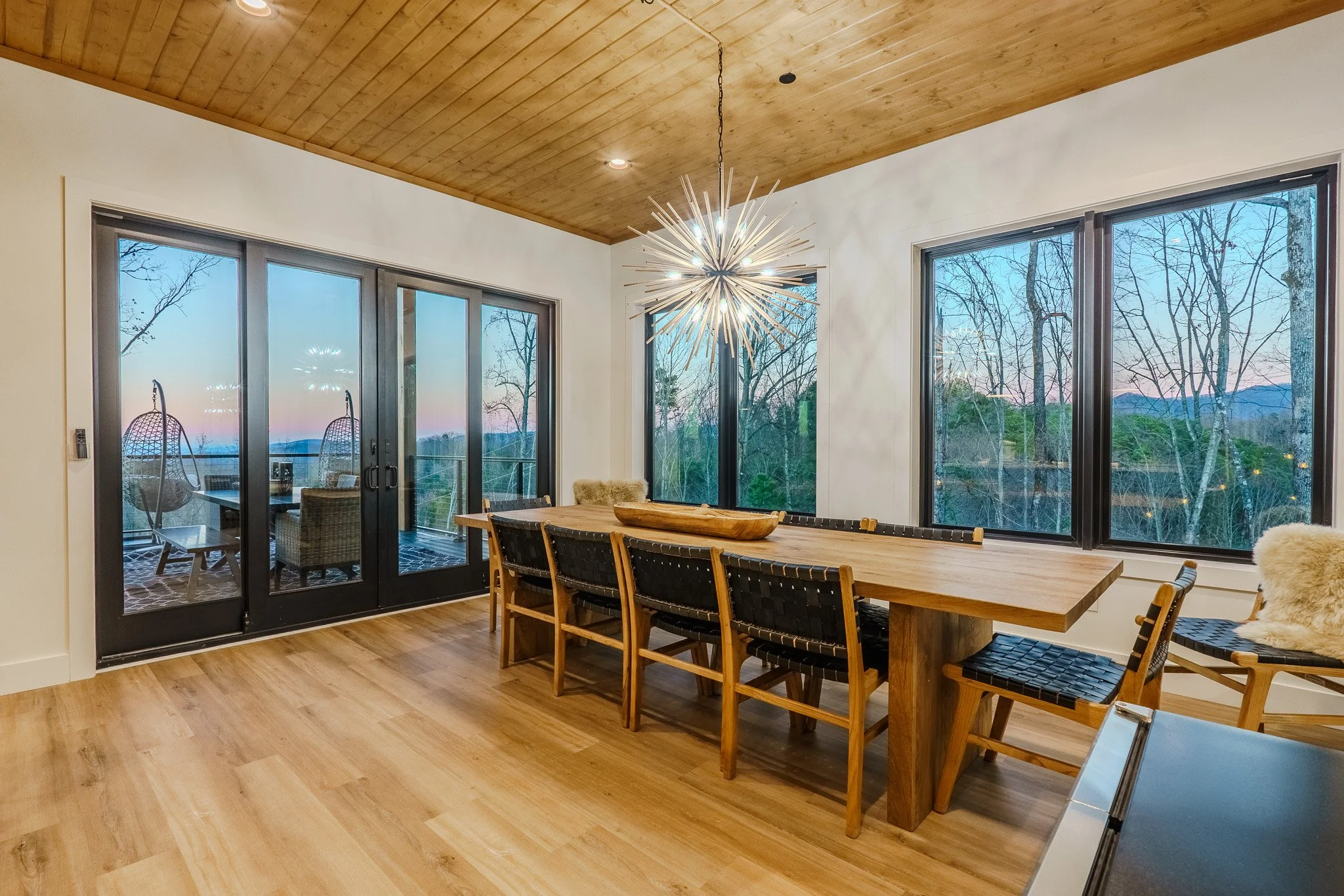 Modern dining room with a large wooden table, seven chairs, ceiling with wood paneling, and a chandelier. Large windows and sliding glass doors reveal a patio and trees outside at sunset.