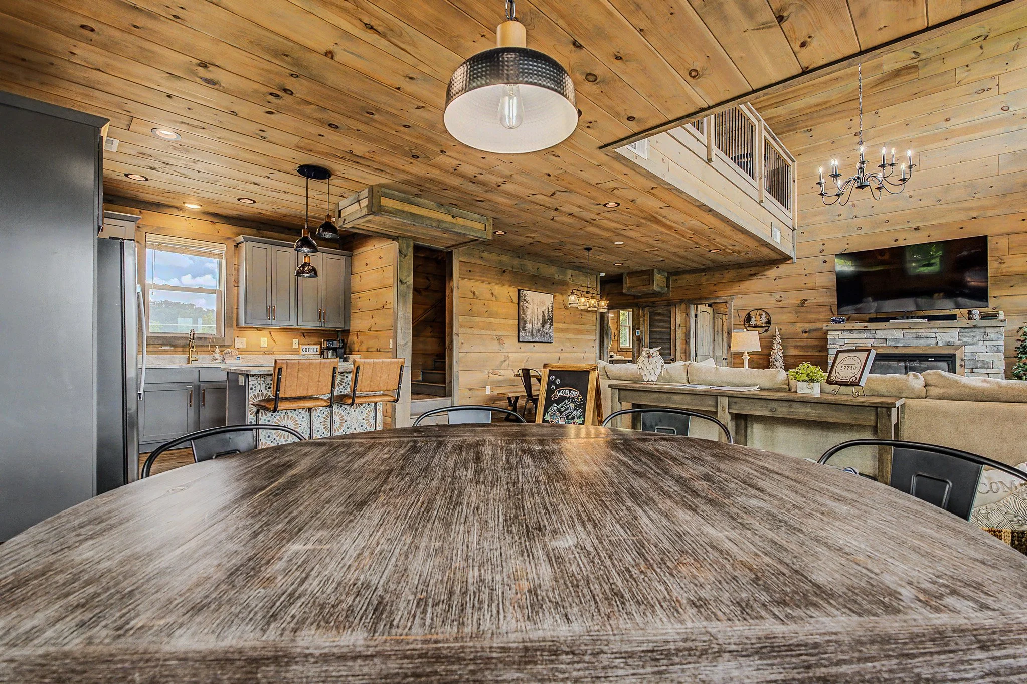 Interior of a rustic wooden house with a dining table in the foreground, kitchen area with gray cabinets, and a living room with a television and chandelier in the background.