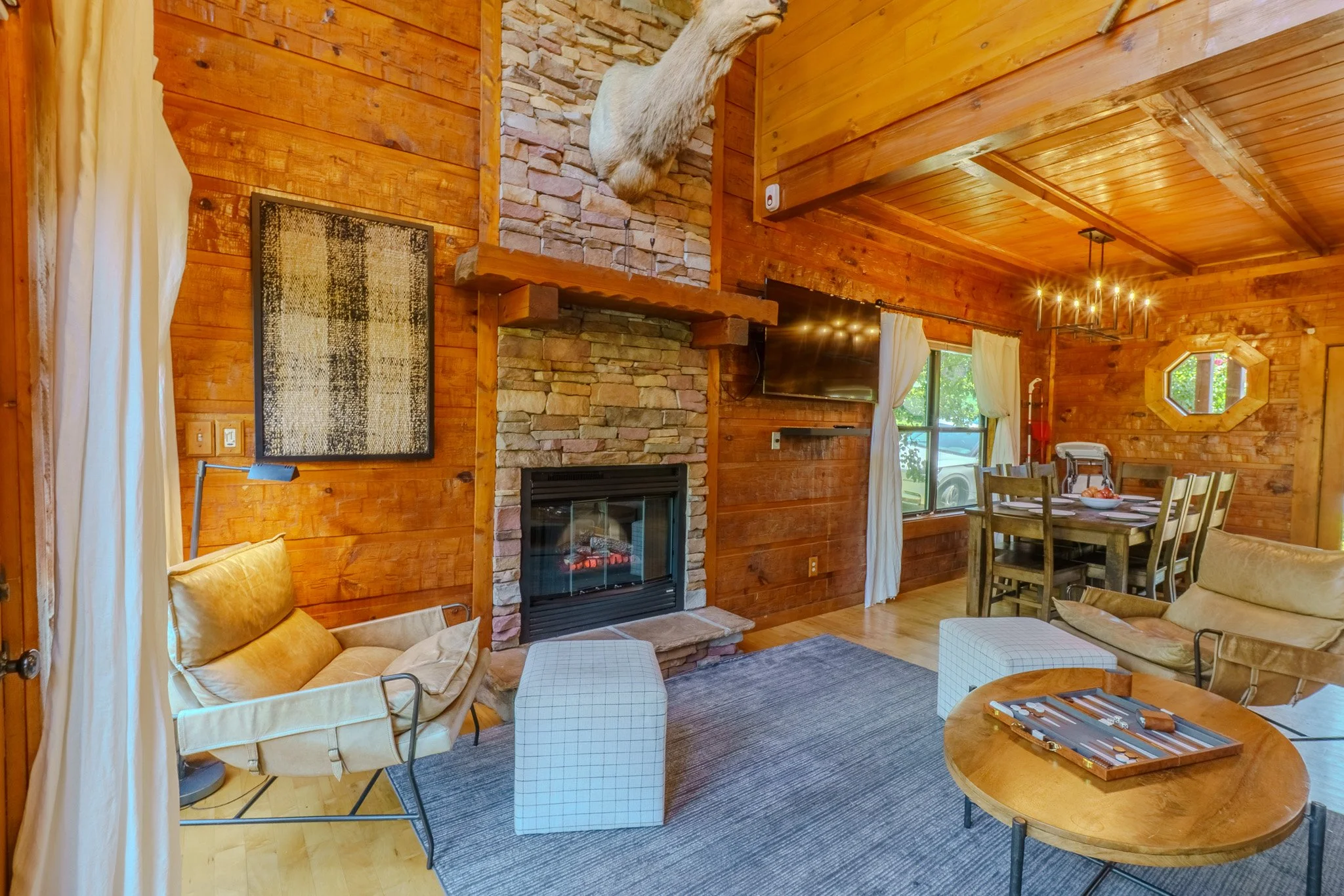 Cozy living room with wood-paneled walls, a fireplace with a stone surround, a mounted animal skull, a flat-screen TV, and a seating area with chairs, ottomans, and a coffee table. There's also a dining area with a table and chairs, a window with cur