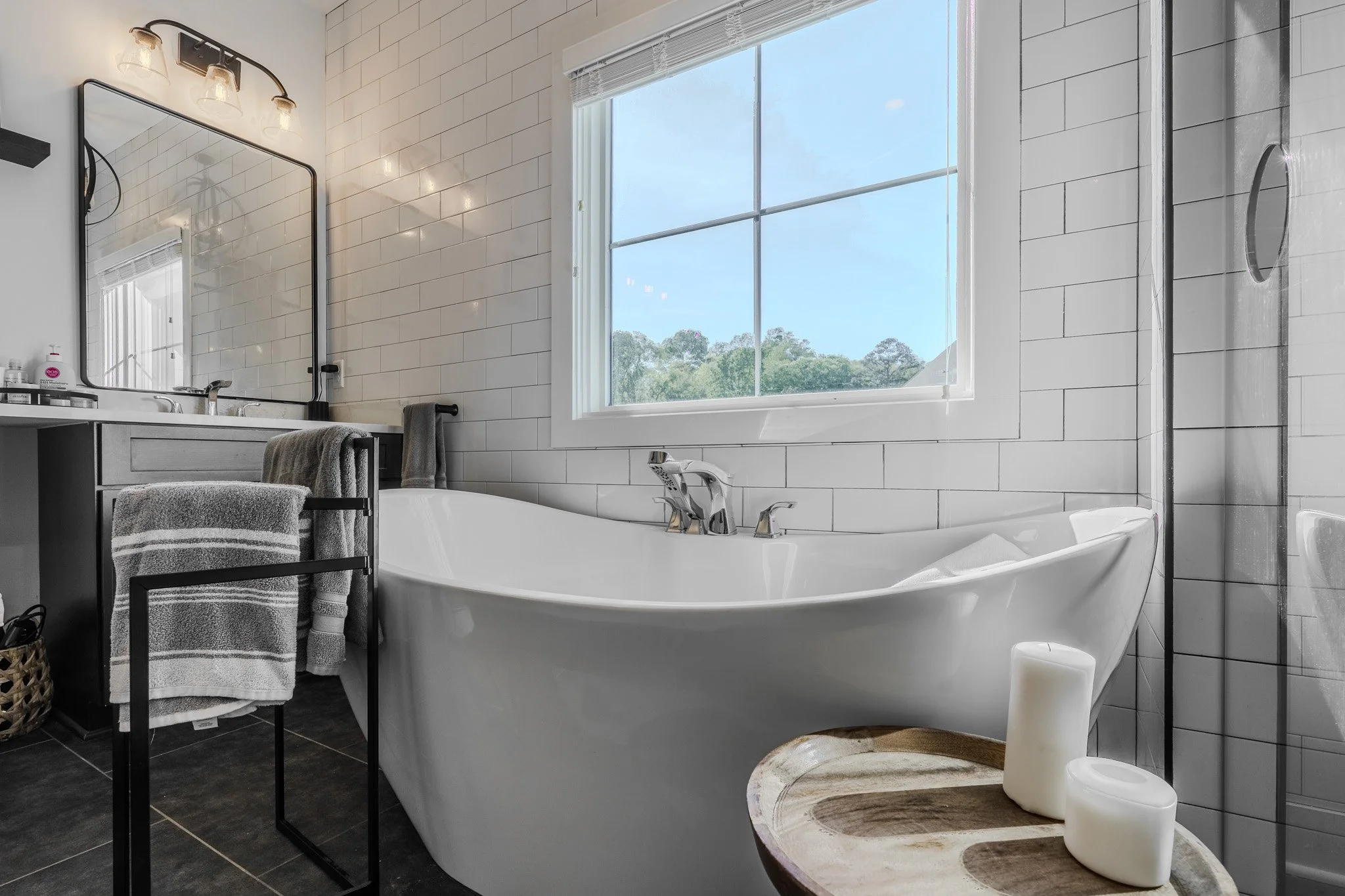 Modern bathroom with a white bathtub, black framed mirror, and gray vanity with towels. Natural light from a large window, white tiled walls, and a shower area with glass door.