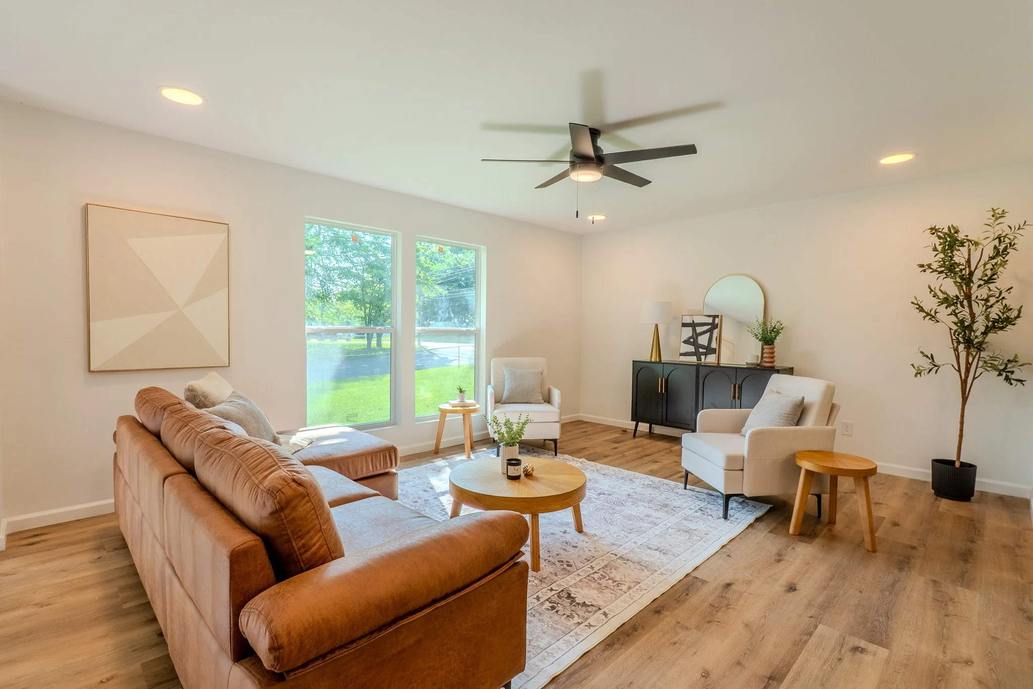 Bright living room with wooden flooring, beige sofa, white armchairs, black sideboard, large window with greenery outside, ceiling fan, and minimal decor.