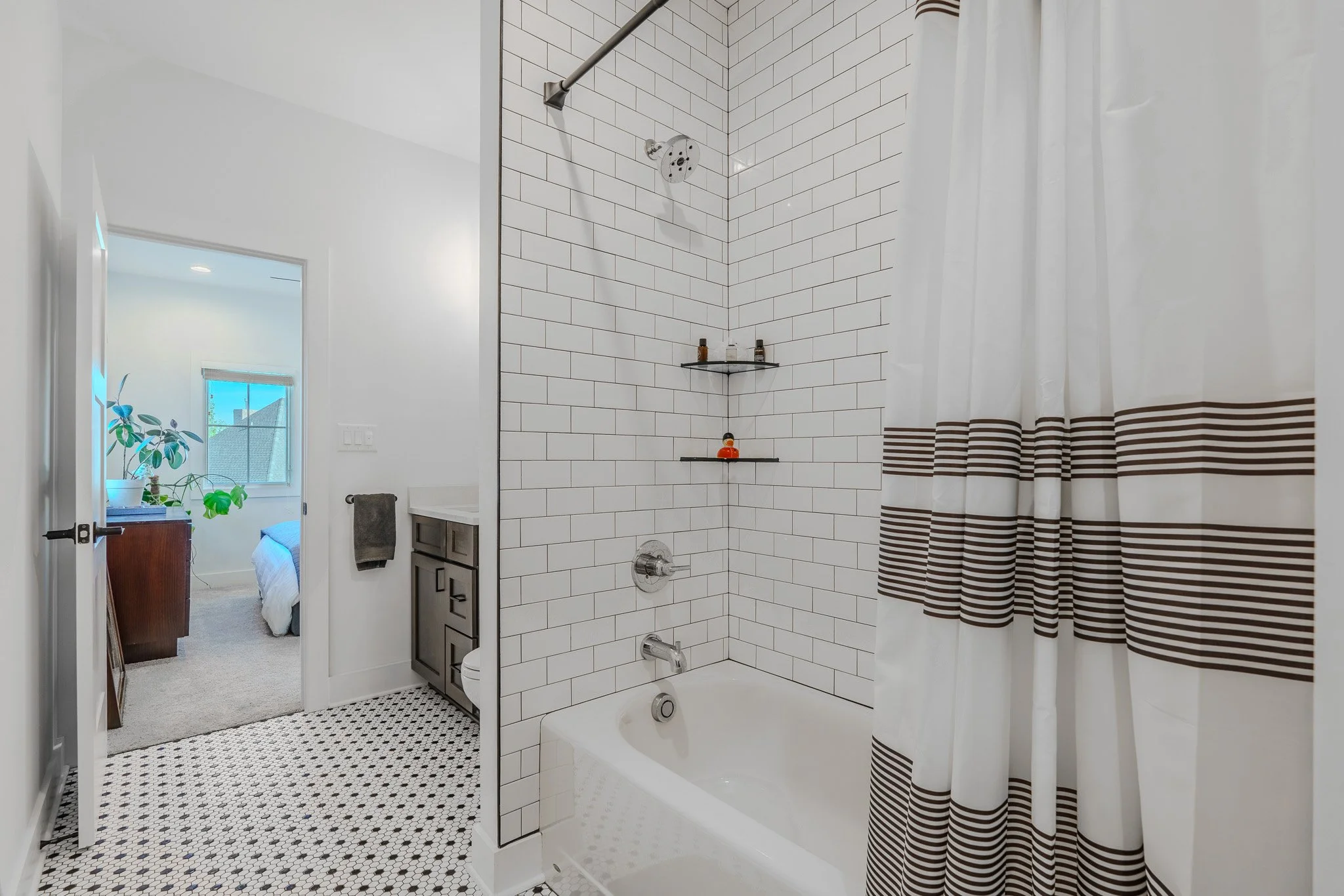 Bathroom with white subway tile walls and a bathtub, a shower curtain with brown stripes, and a view into a bedroom with a bed, window, and a plant.