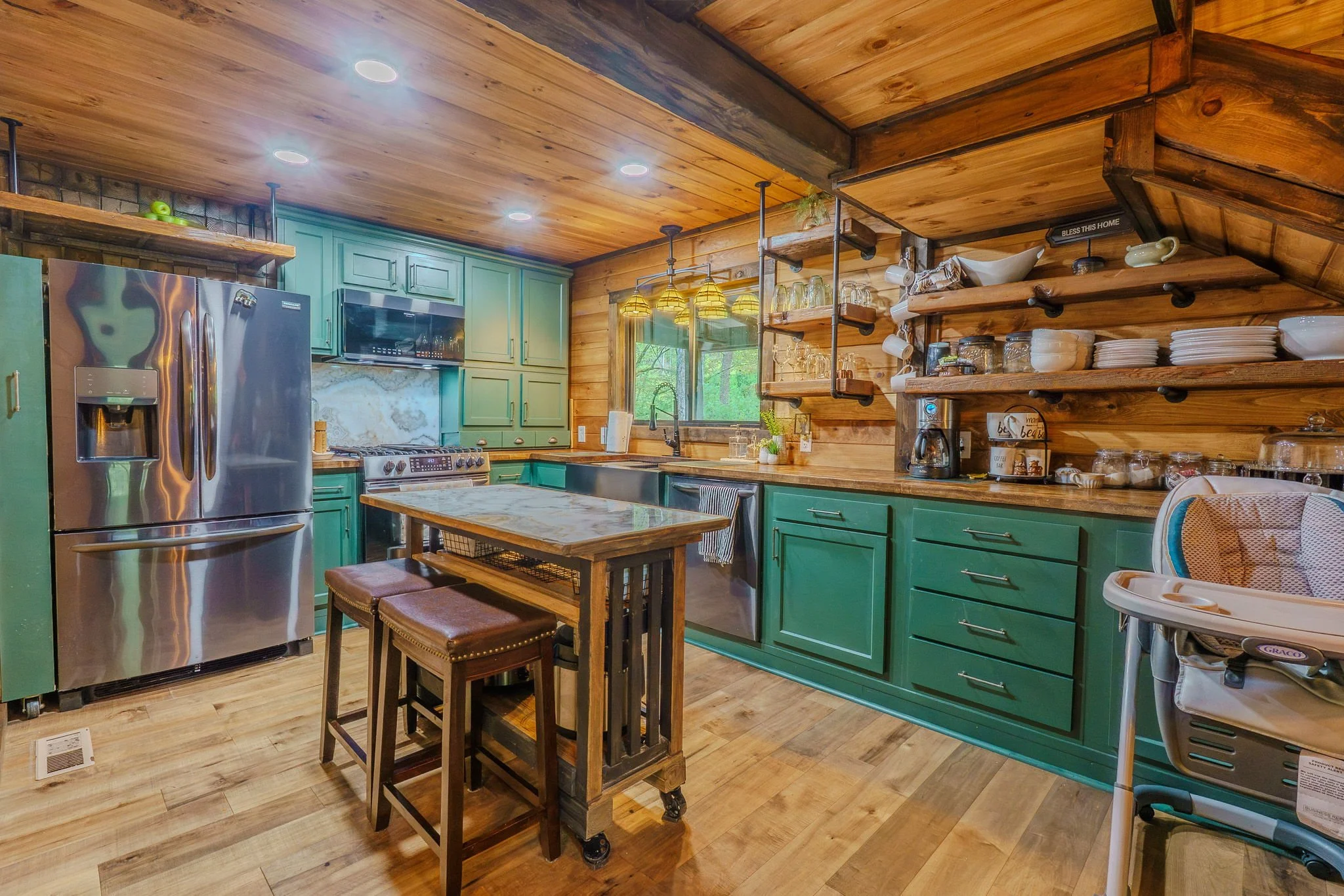 Rustic kitchen with green cabinets, wood-paneled ceiling and walls, stainless steel refrigerator, and open wooden shelves displaying dishes and glassware.