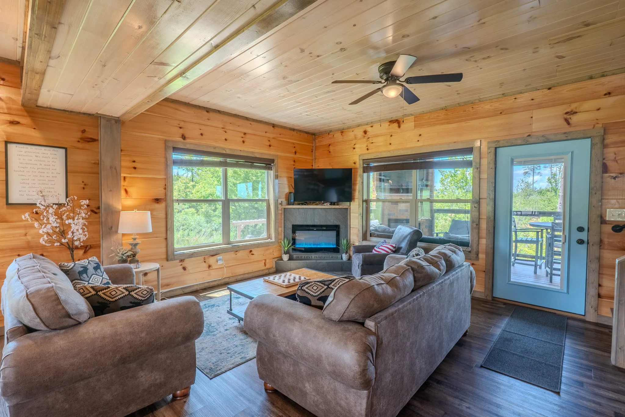 Living room with wood-paneled walls and ceiling, featuring a ceiling fan, a flat-screen TV above a fireplace, a sofa, armchairs, a coffee table, and windows showing green trees outside.