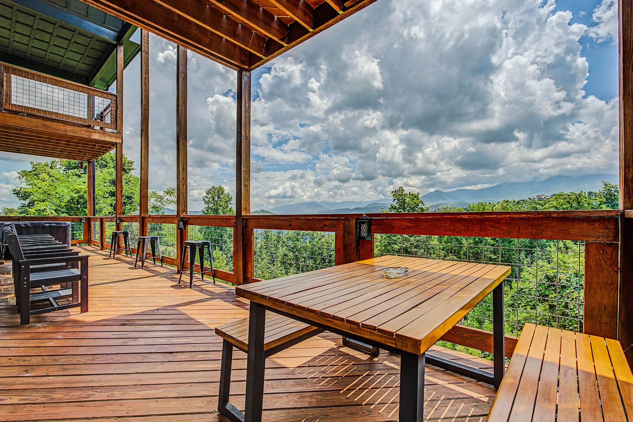 Wooden balcony with tables and chairs overlooking a green landscape and distant mountains, under cloudy sky.