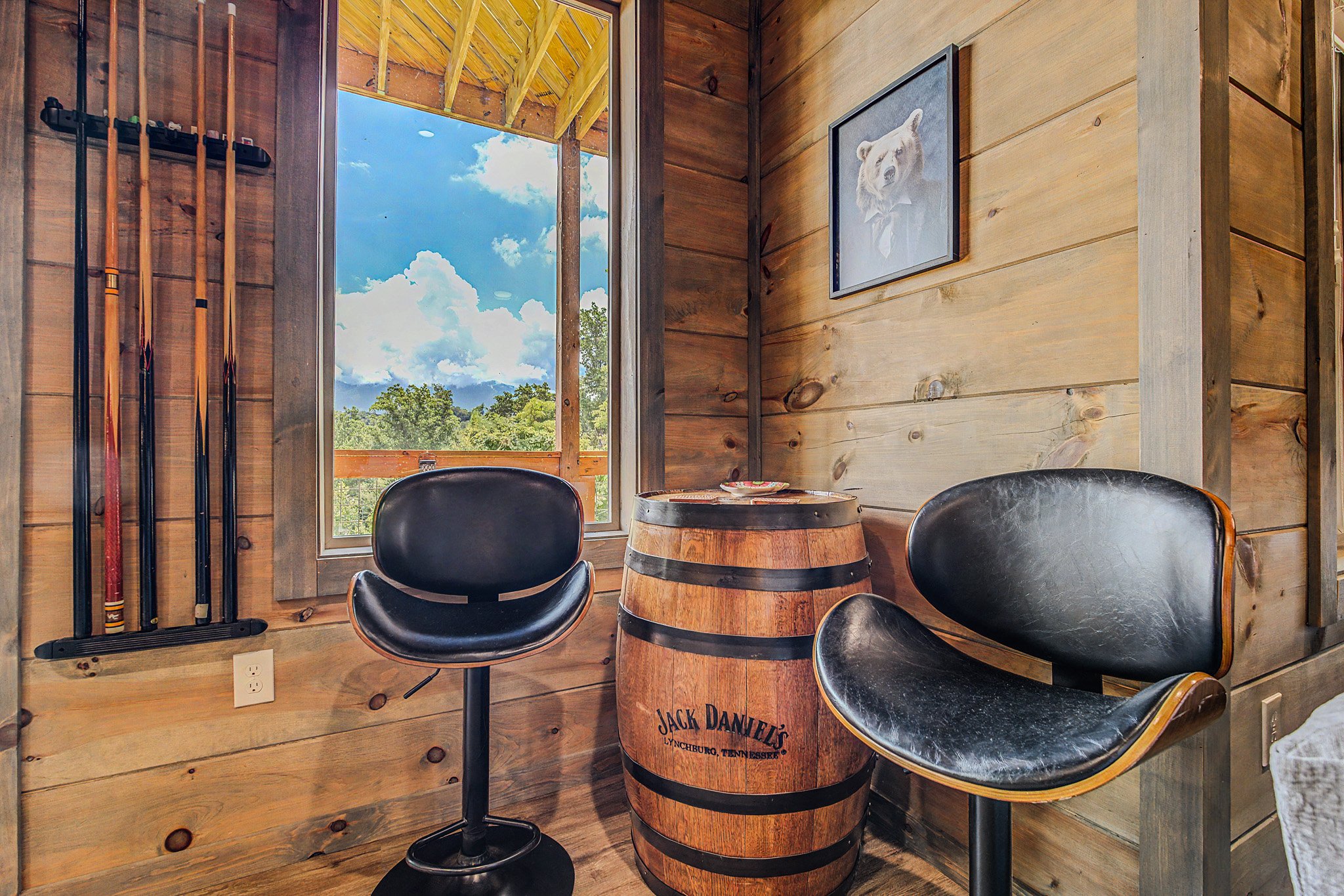Rustic room with wooden walls, a window showing trees and a blue sky with clouds, and a small seating area featuring a barrel-style table with Jack Daniel's branding and two black chairs.