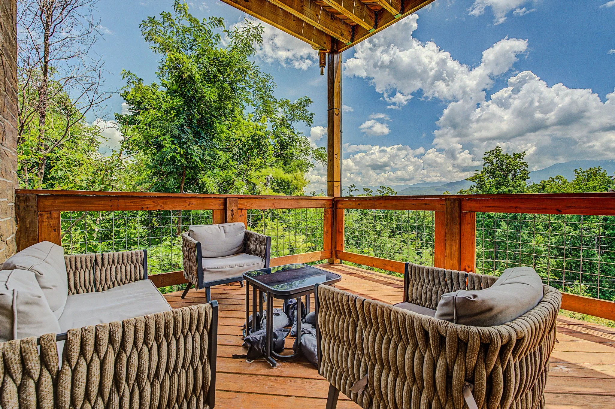 A wooden deck with outdoor seating including cushioned chairs and a small table, overlooking a lush green landscape with trees and mountains in the distance under a partly cloudy sky.