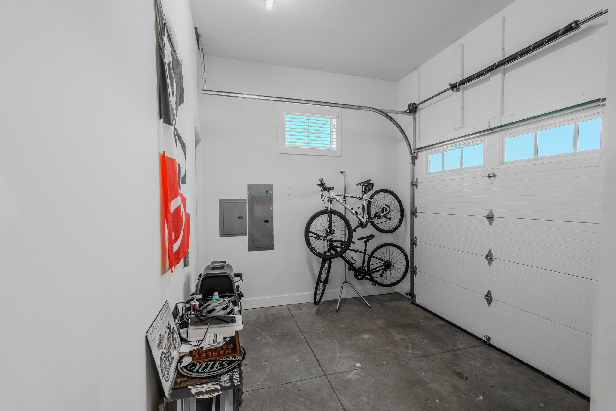 A garage with a white roll-up door, two bicycles mounted on a wall, a small window, a workbench with tools, and various storage boxes.