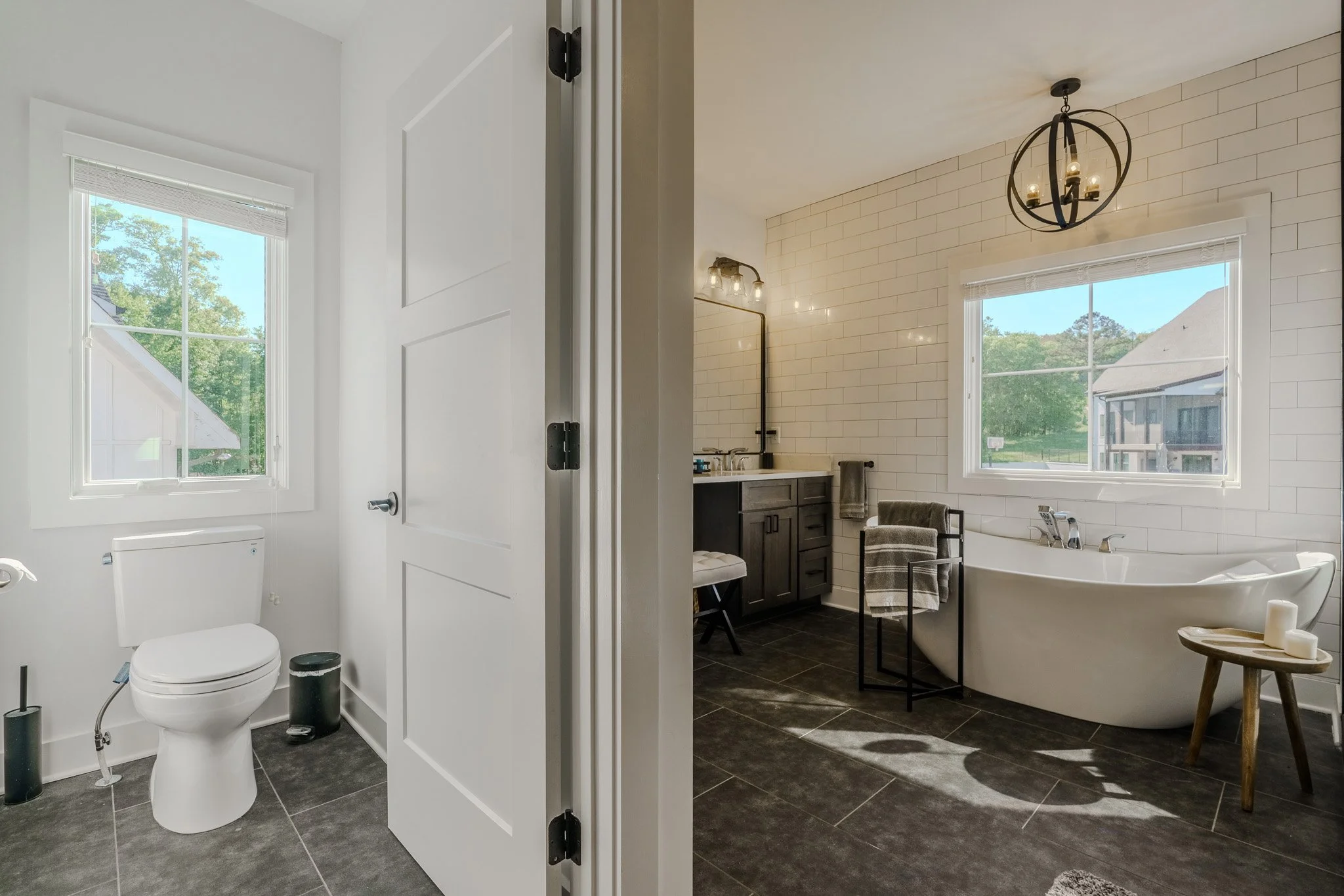 A bathroom with a white toilet, black floor tiles, and a window showing trees outside. Adjacent to it is a larger bathroom with a bathtub near a window, a dark vanity with a mirror, and ceiling light fixtures. The rooms are separated by a door.