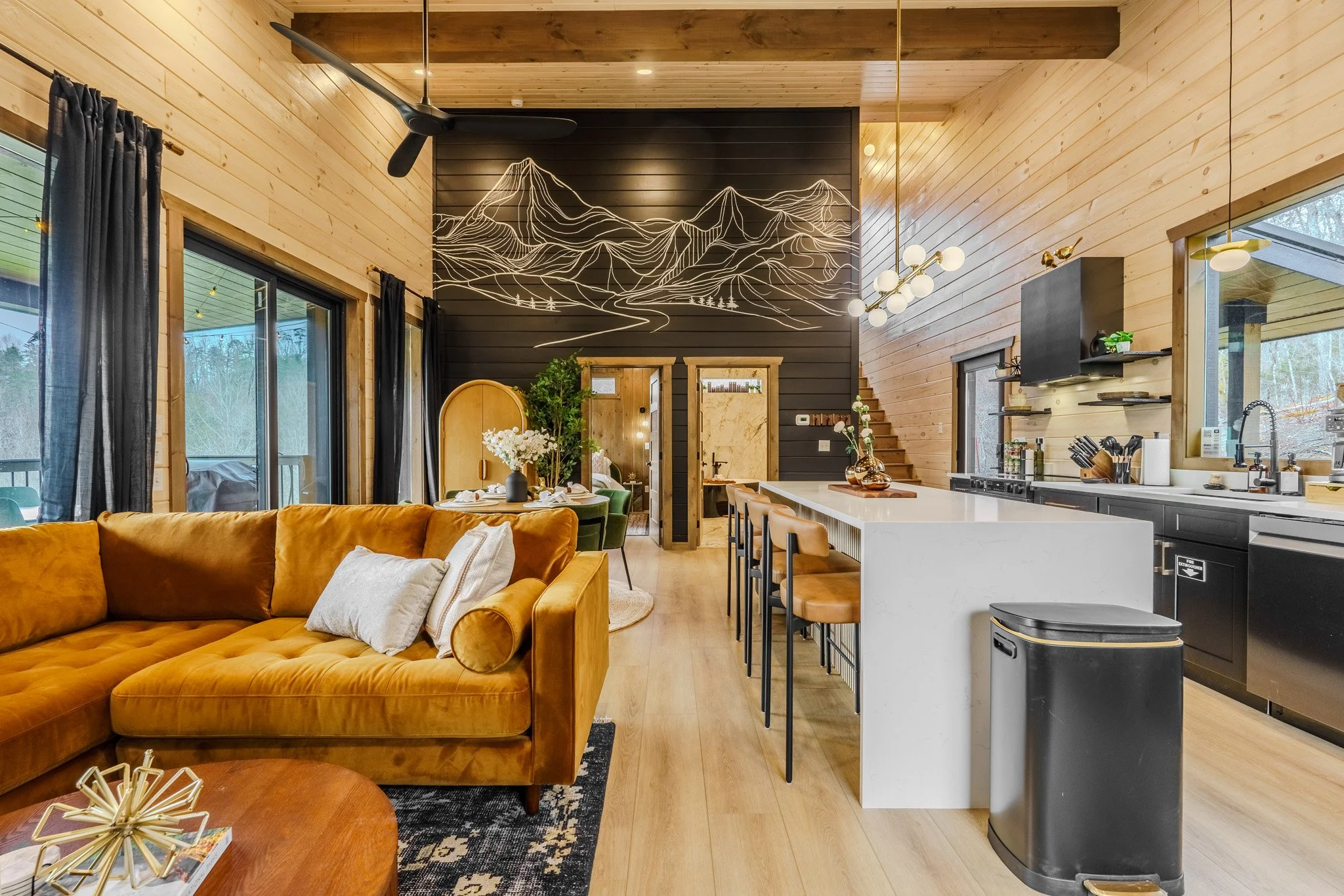 Open-concept living room and kitchen with wooden walls, black and white accents, and modern furniture, including a mustard sofa, bar stools, and chandelier lights.