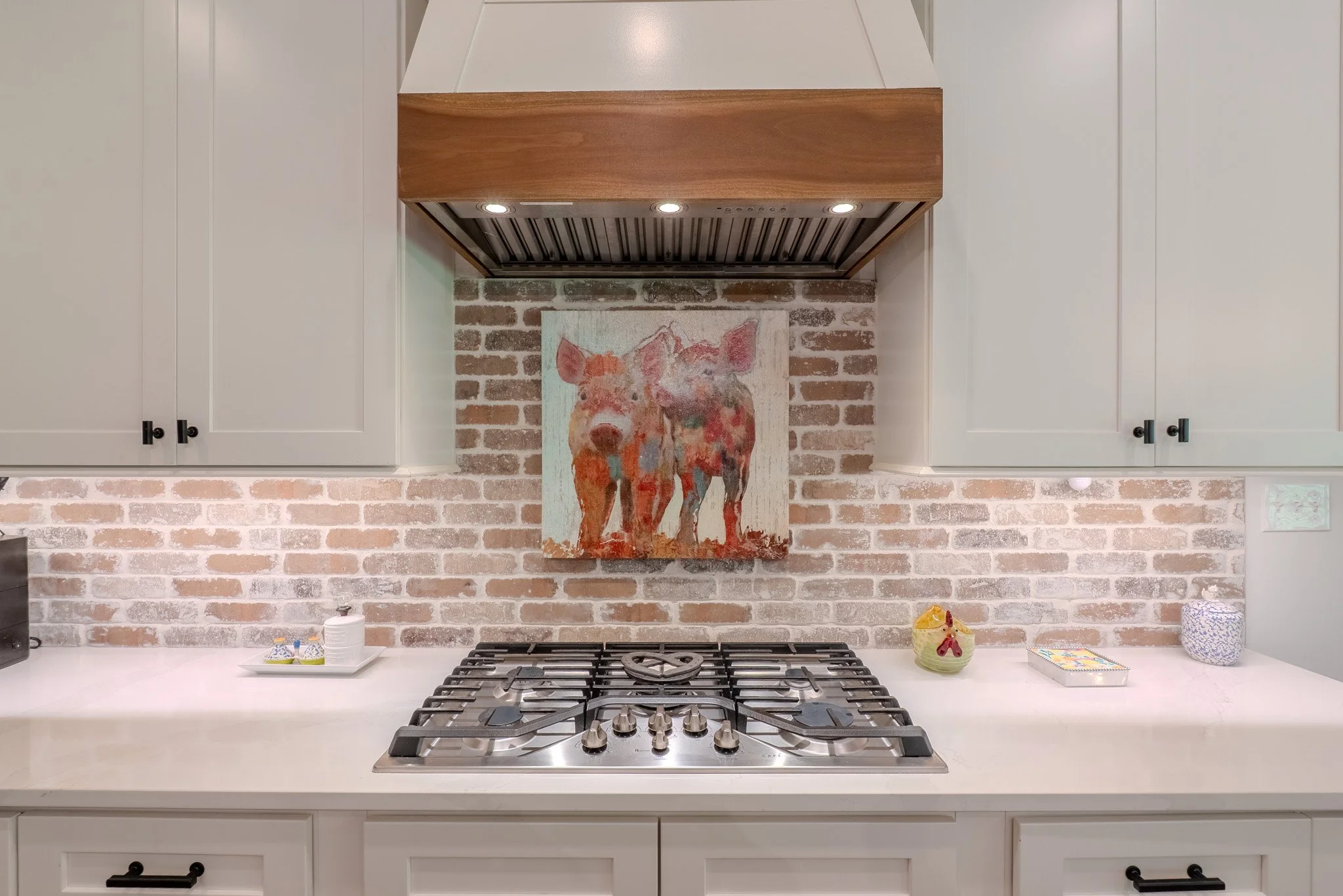 Kitchen with white cabinets, a brick backsplash, and a painting of two piglets hanging above a stainless steel gas stove.