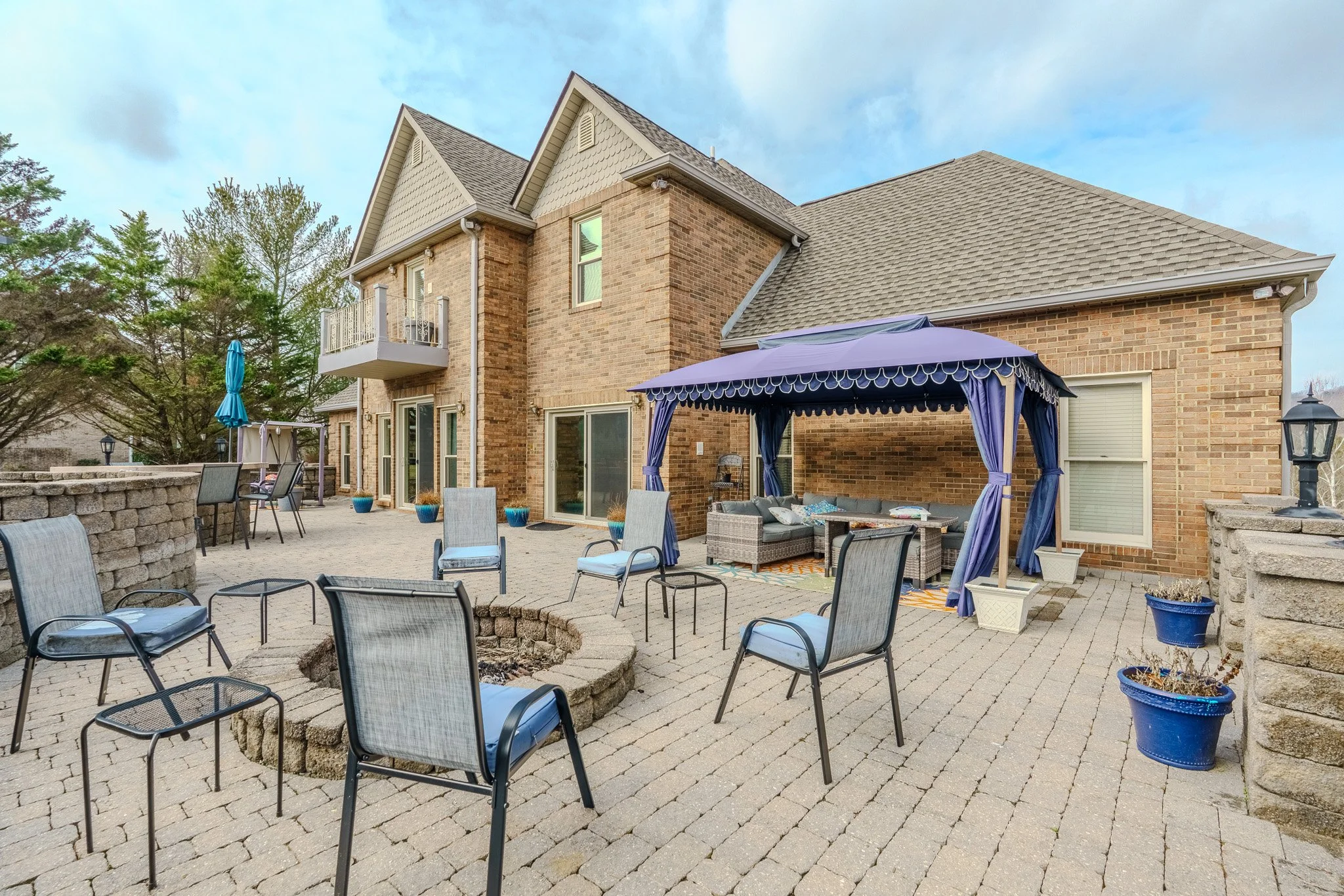 Backyard patio with outdoor seating including chairs, a gazebo with curtains and cushions, potted plants, and a fire pit, surrounded by a brick house and stone wall.