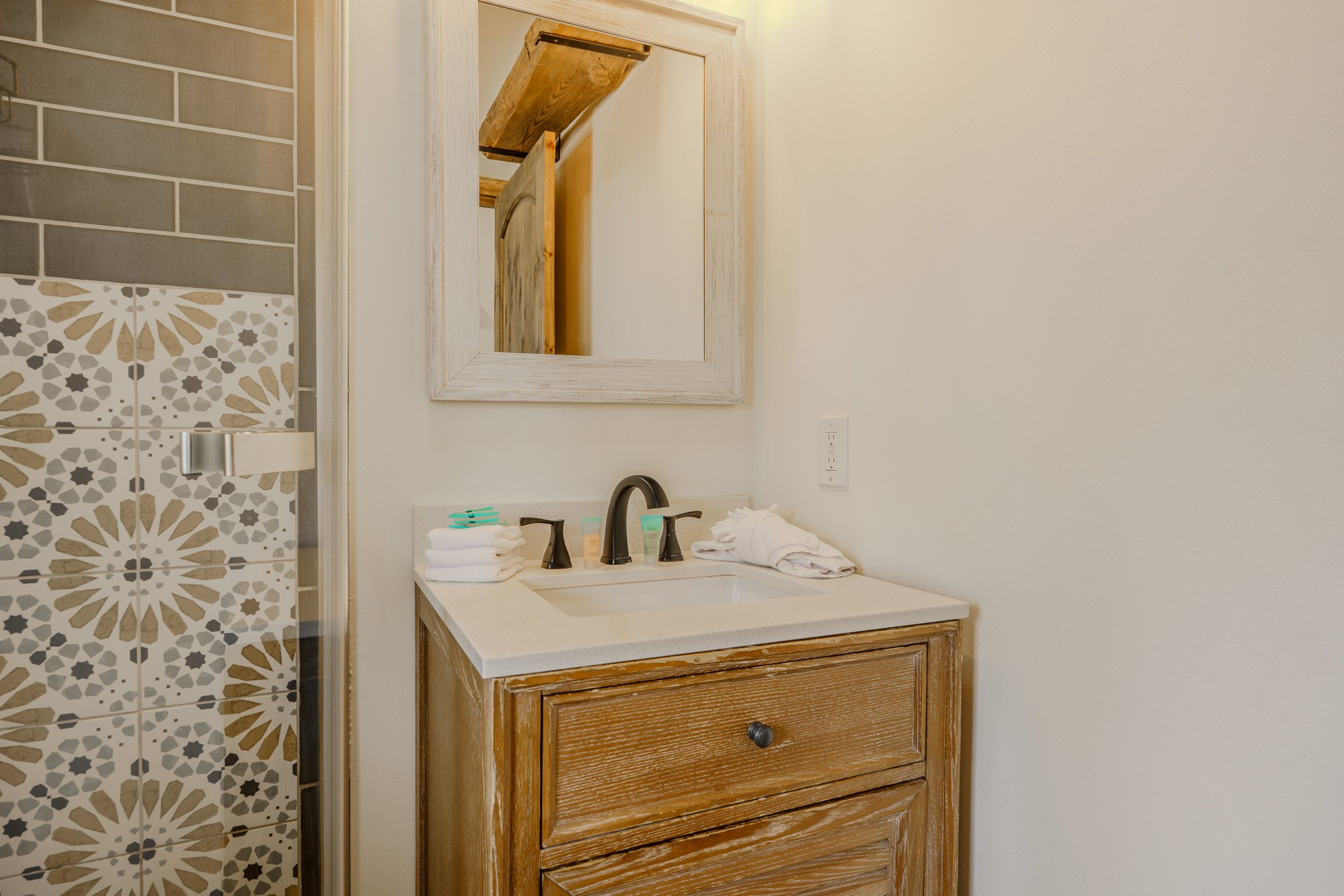 Bathroom vanity with a wooden cabinet, white countertop, black faucet, mirror, and toiletries, alongside a partly visible tiled shower with patterned tiles.