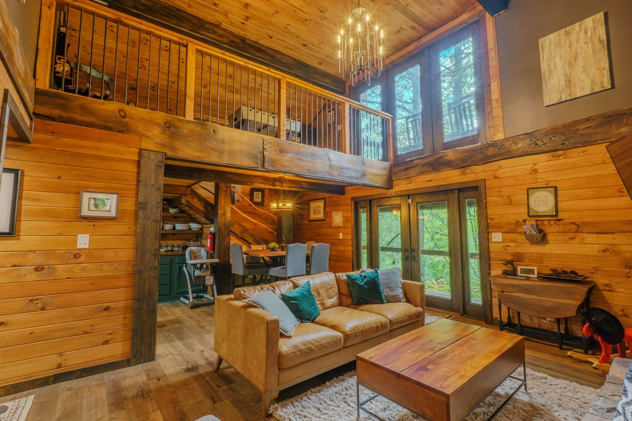 Interior view of a rustic wooden cabin living room with a second-story loft. Features include a tan leather sofa with teal pillows, a wooden coffee table, large windows with a forest view, and wooden walls and ceiling. There is a chandelier overhead 