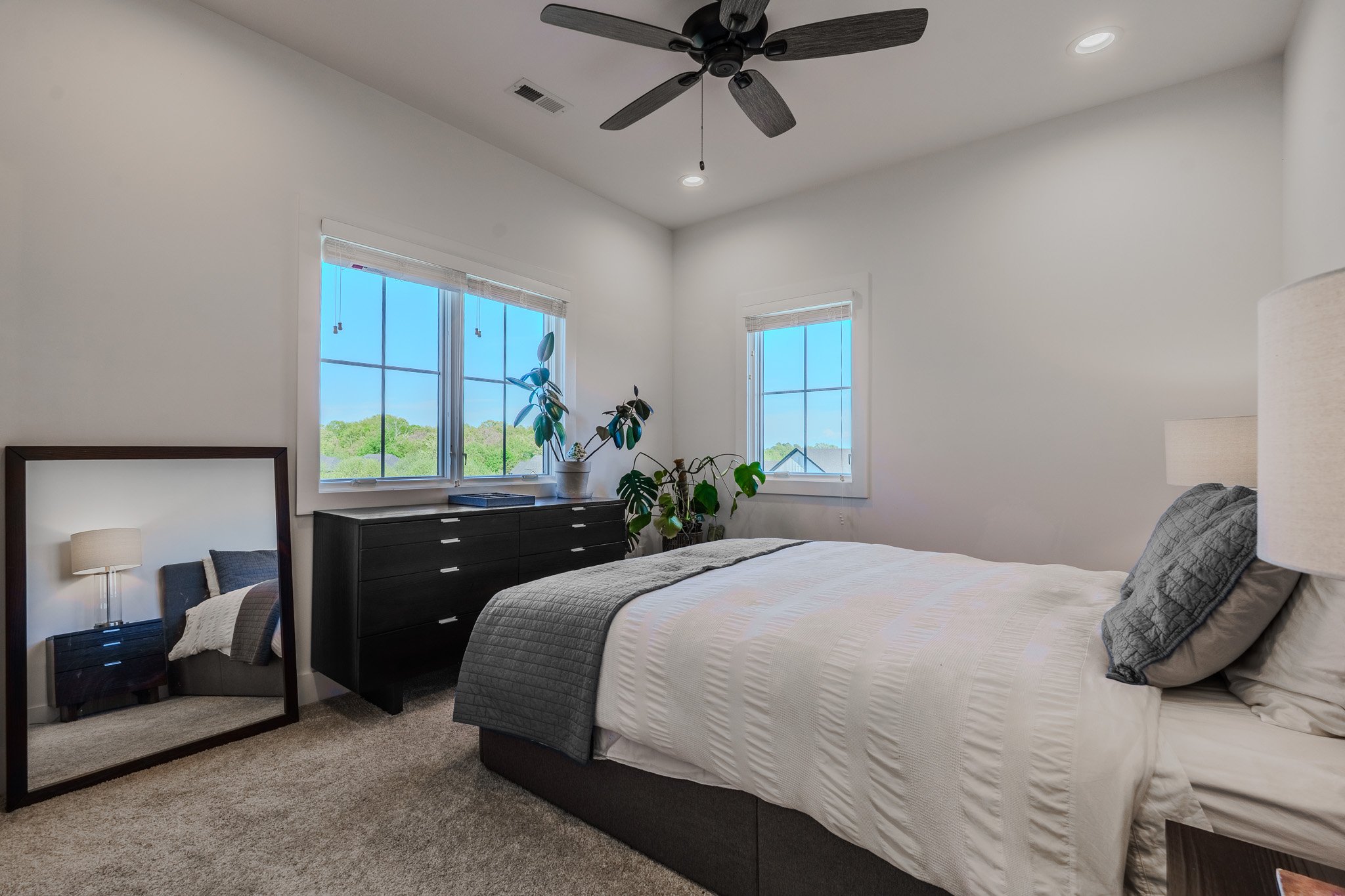 A modern bedroom with white walls, light beige carpet, a bed with white bedding and gray pillows, two large windows with blinds, a black dresser, and a full-length mirror leaning against the wall reflecting a nightstand with a lamp and bed.