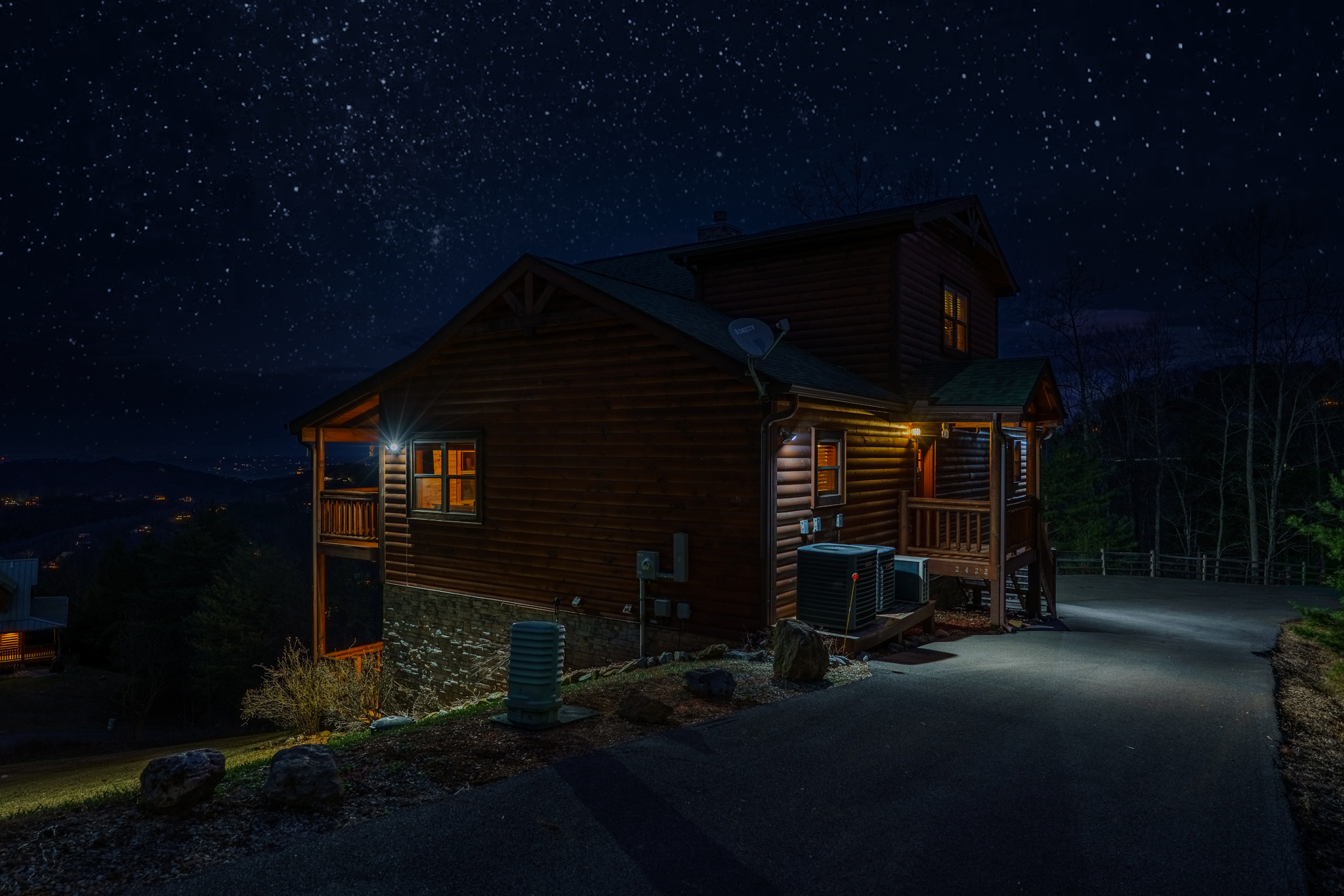 A wooden house on a hill at night with illuminated windows and exterior lights, a starry sky overhead, and trees in the background.