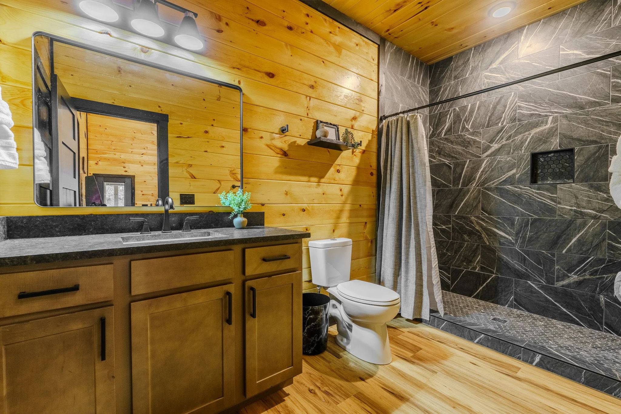 Bathroom with wooden walls and flooring, a black countertop with a sink, a large mirror, a small potted plant, a toilet, and a walk-in shower with dark marble walls and a window.