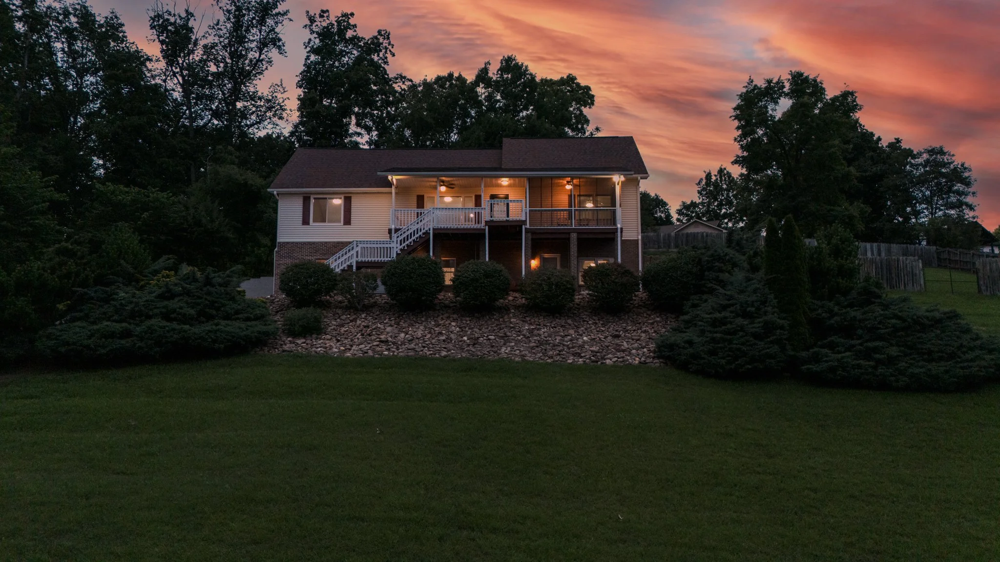 A house on a hill at sunset with outdoor lights on, surrounded by trees and shrubs, with a grassy lawn in the foreground.