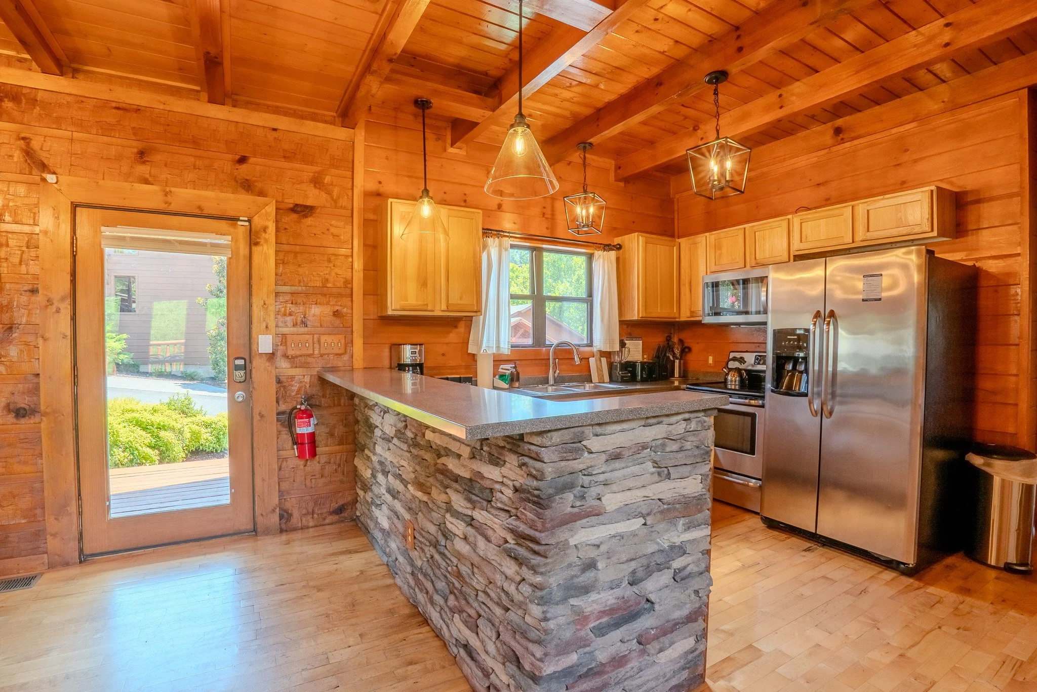 Wooden kitchen with stone island, stainless steel fridge, microwave, and wooden cabinets, illuminated by hanging pendant lights, with a window above the sink and a glass door leading outside.