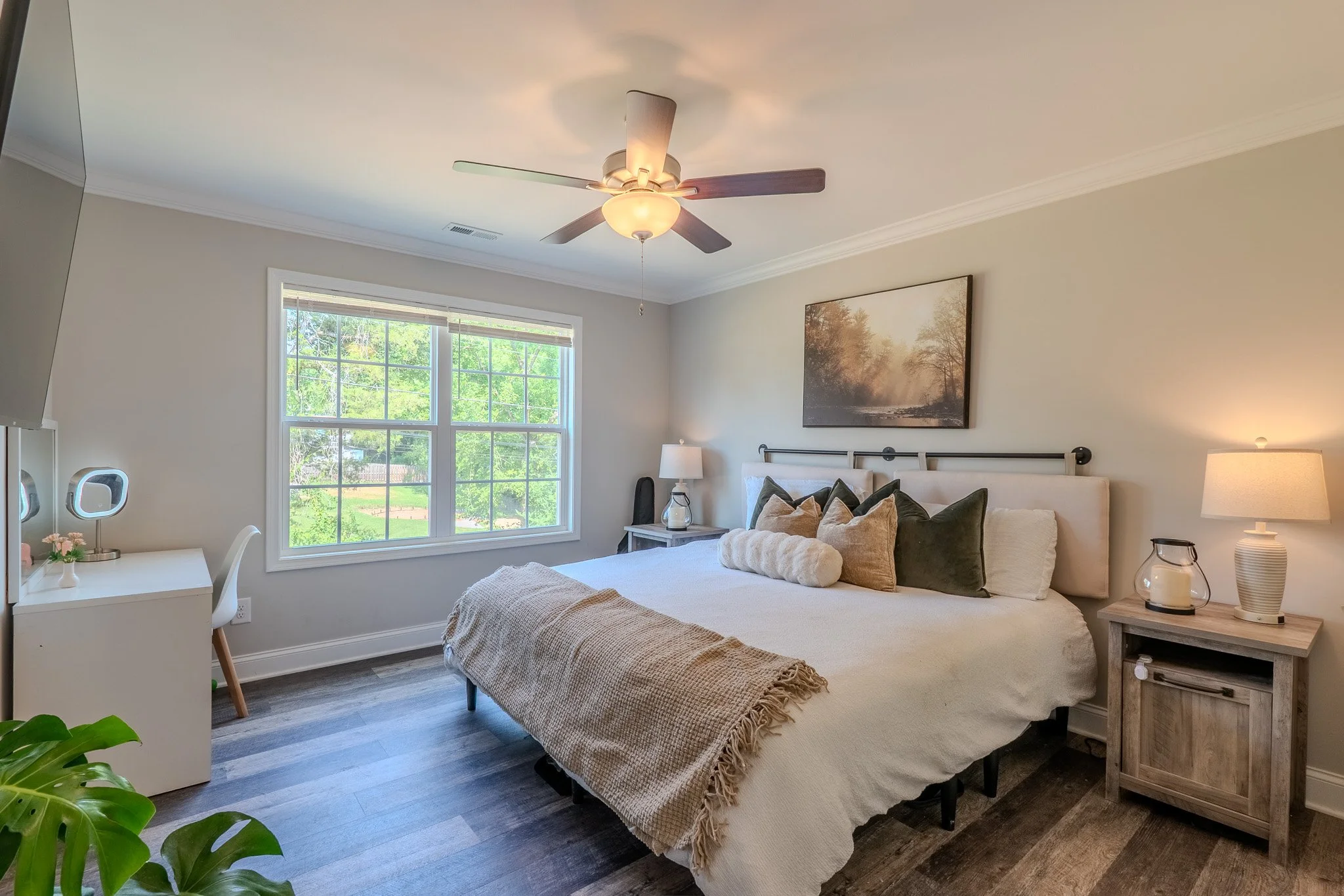 A neatly made bedroom with a large bed, multiple pillows, a nightstand with a lamp, a window showing trees outside, and a ceiling fan.