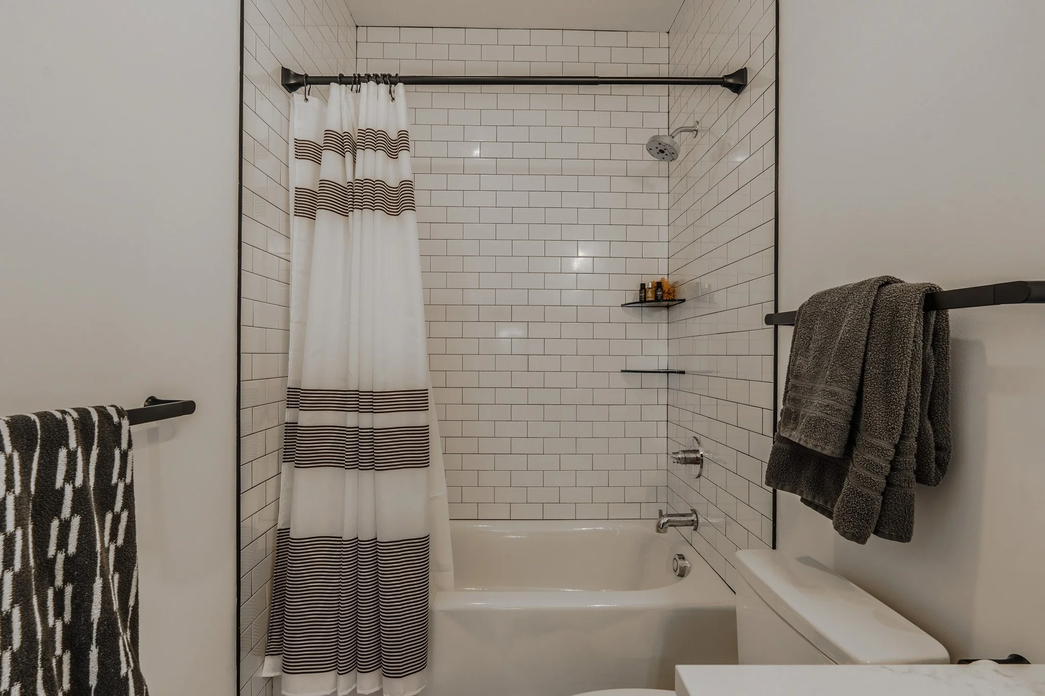 Bathroom with a bathtub and shower area enclosed by a white curtain with dark horizontal stripes, white tiled walls with dark grout, black towel bars with towels, and a small corner shelf with toiletries.
