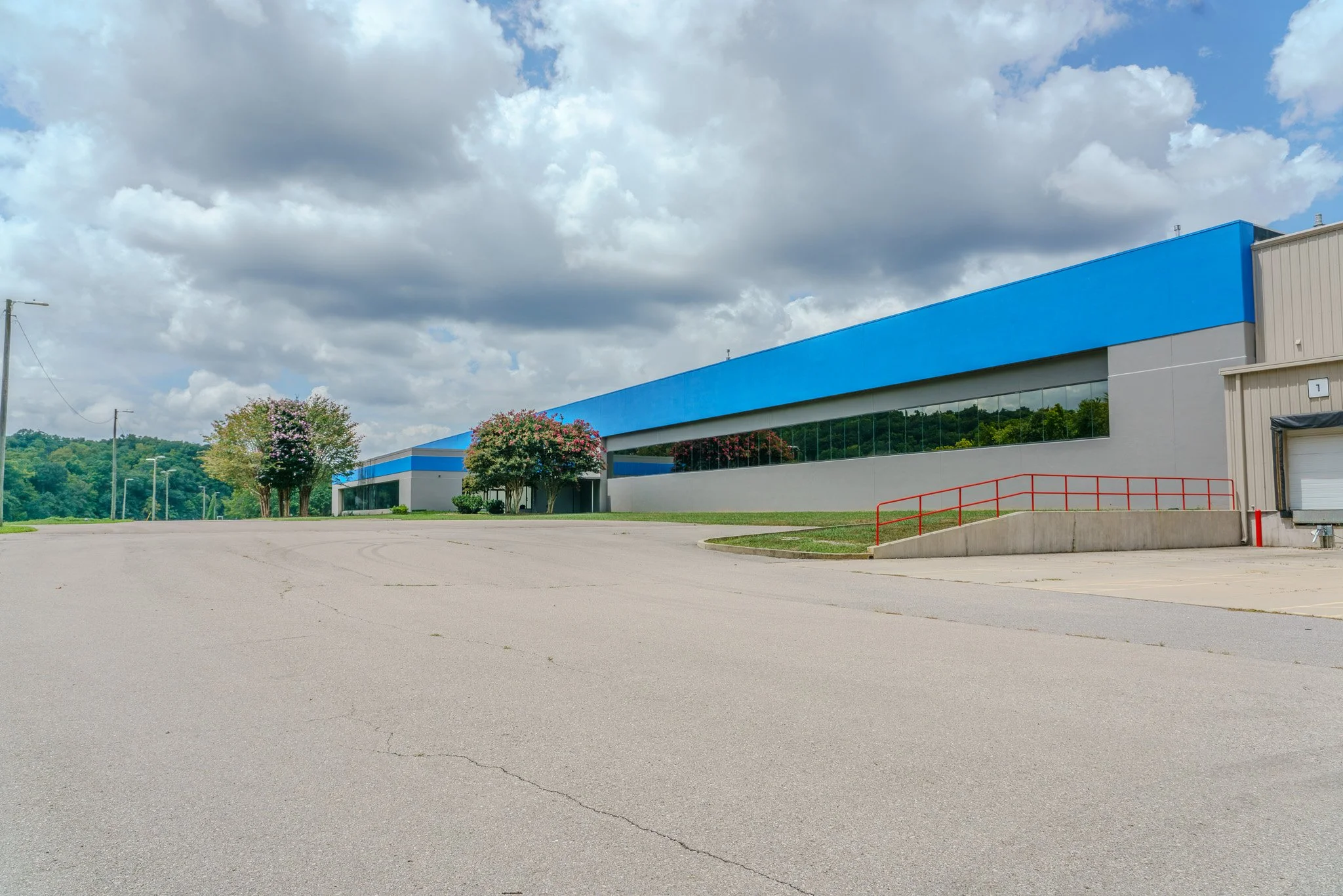 An empty parking lot in front of a large industrial building with a blue roof and reflective windows, surrounded by green trees and cloudy sky.