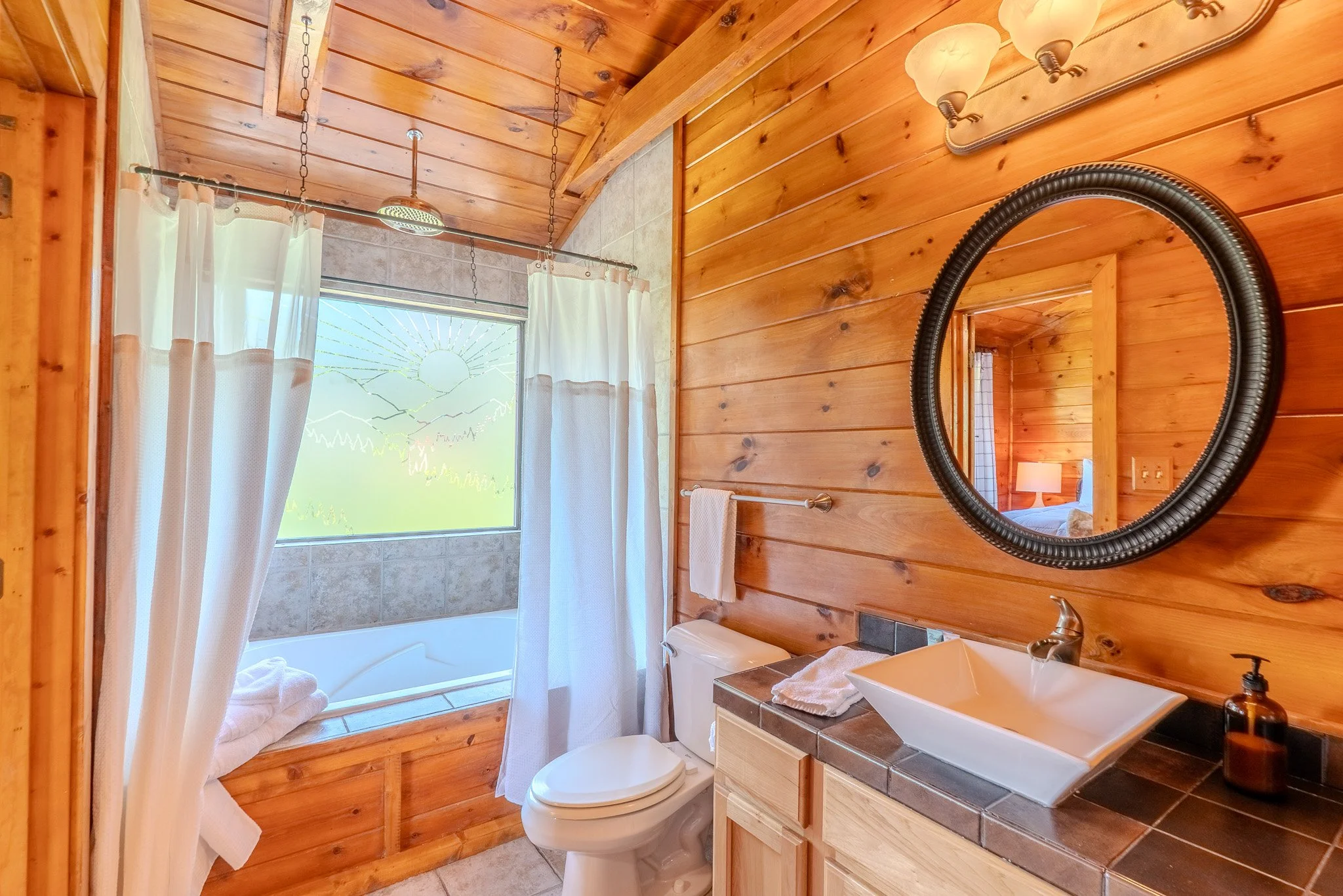 Bathroom with wooden walls, a bathtub with a window above it, a toilet, and a sink with a mirror. Curtains hang in front of the window, and there is a towel on a rack. The reflection shows a bedroom with a lamp.