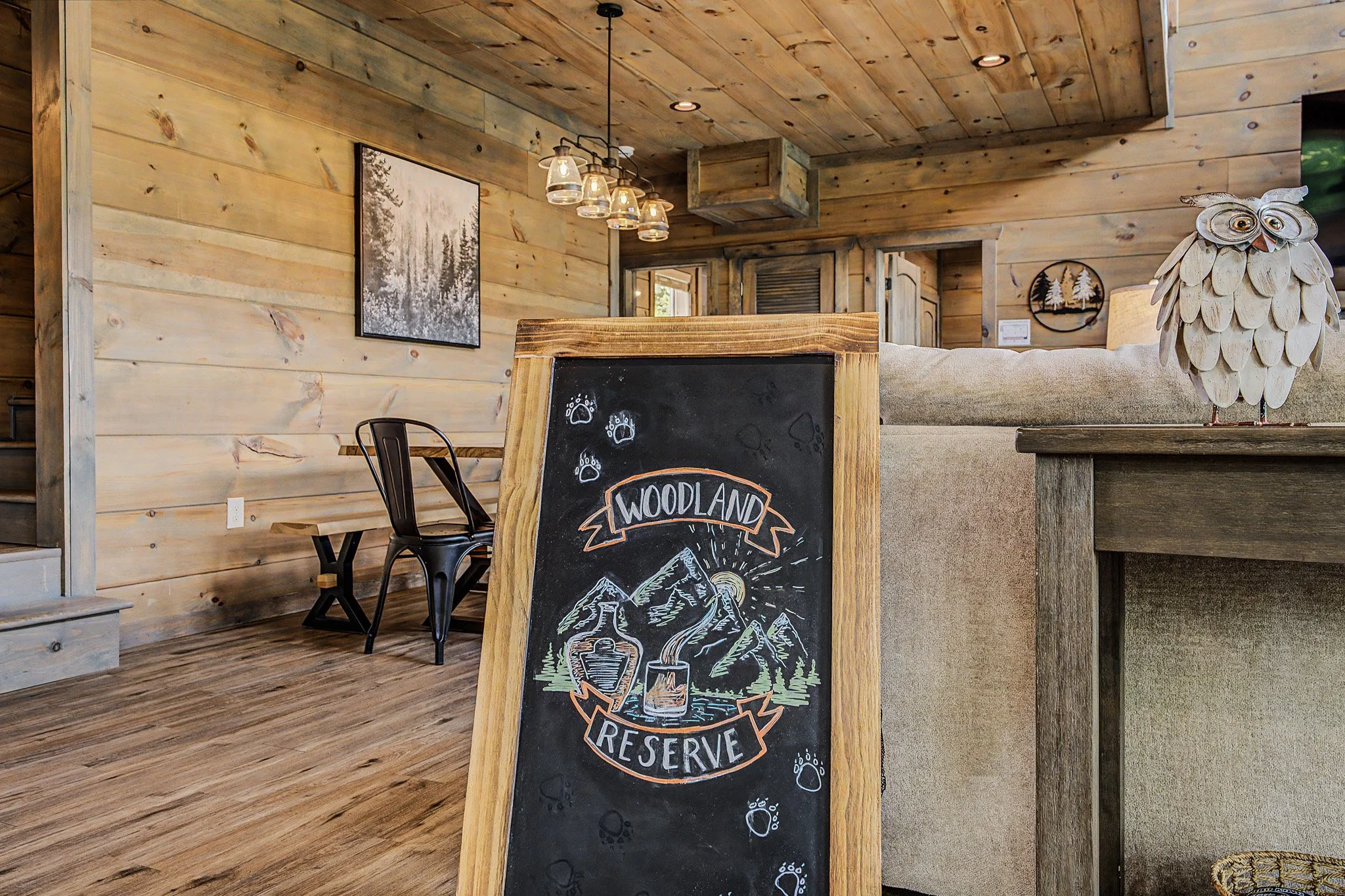 Interior of a rustic wooden room with a chalkboard displaying the word 'RESERVE' and mountain scenery, a decorative owl figure on the right, and a hanging light fixture with multiple bulbs.