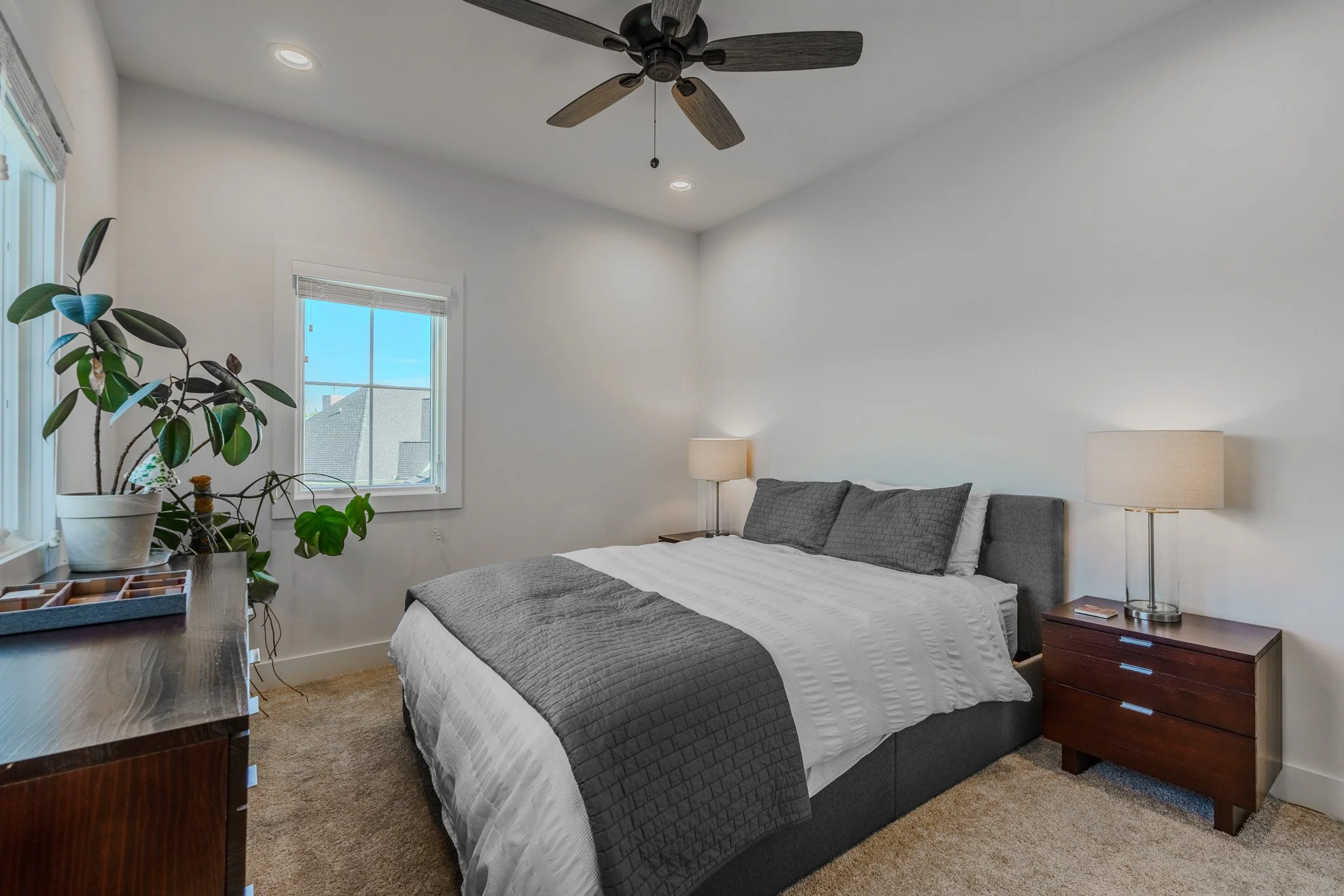 A bedroom with a king-sized bed, gray headboard, white and gray bedding, two bedside lamps, a window with blinds, a plant on a dresser, and a ceiling fan.