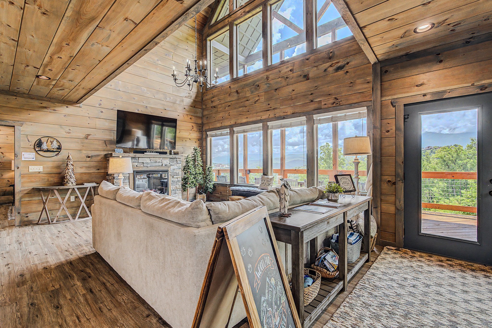 Living room with wooden walls and ceiling, large windows showing a mountain view, beige sofa, fireplace, chandelier, decorative items, and a door to a balcony.
