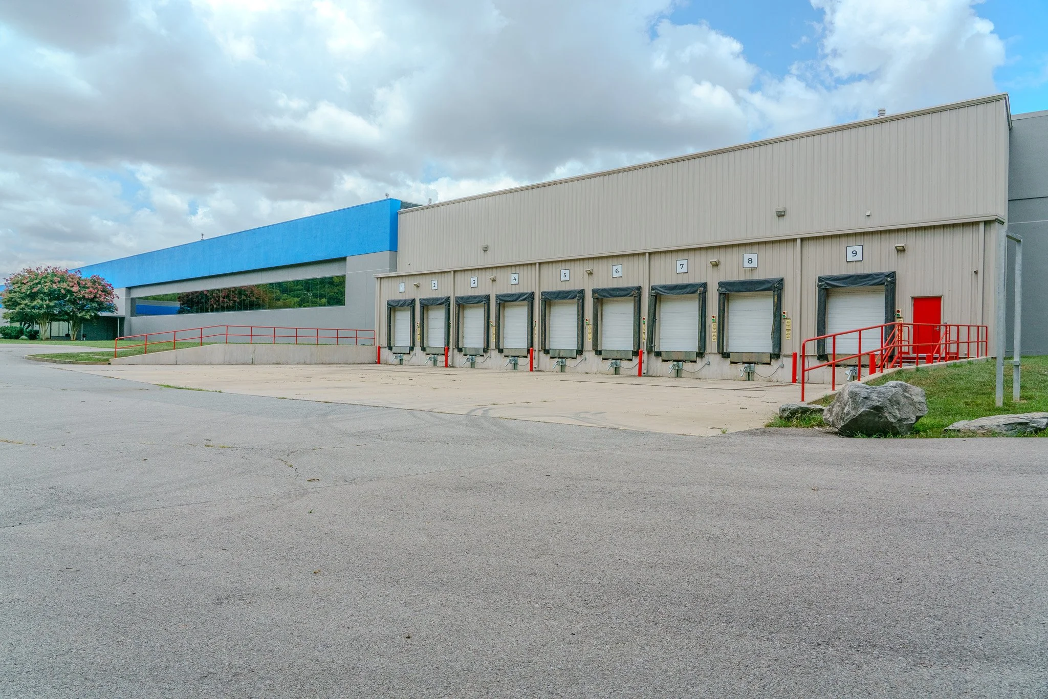 Empty warehouse with multiple loading docks and a red door, surrounded by a paved lot and a grassy area with rocks, under a partly cloudy sky.