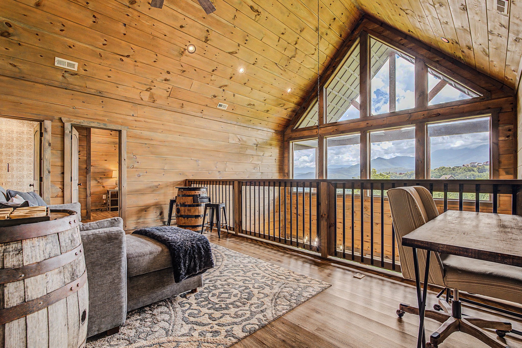 A spacious living room with wood-paneled walls and ceiling, large windows offering mountain and sky views, a gray sofa with black and white pillows, a patterned rug, a dining table with striped chairs, and rustic decor elements such as metal barrels 