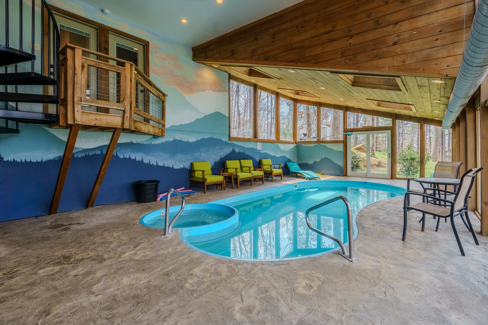 Indoor pool area with a small round hot tub, green chairs along the wall, wooden and glass walls with a mountain mural, and large windows showing trees outside.