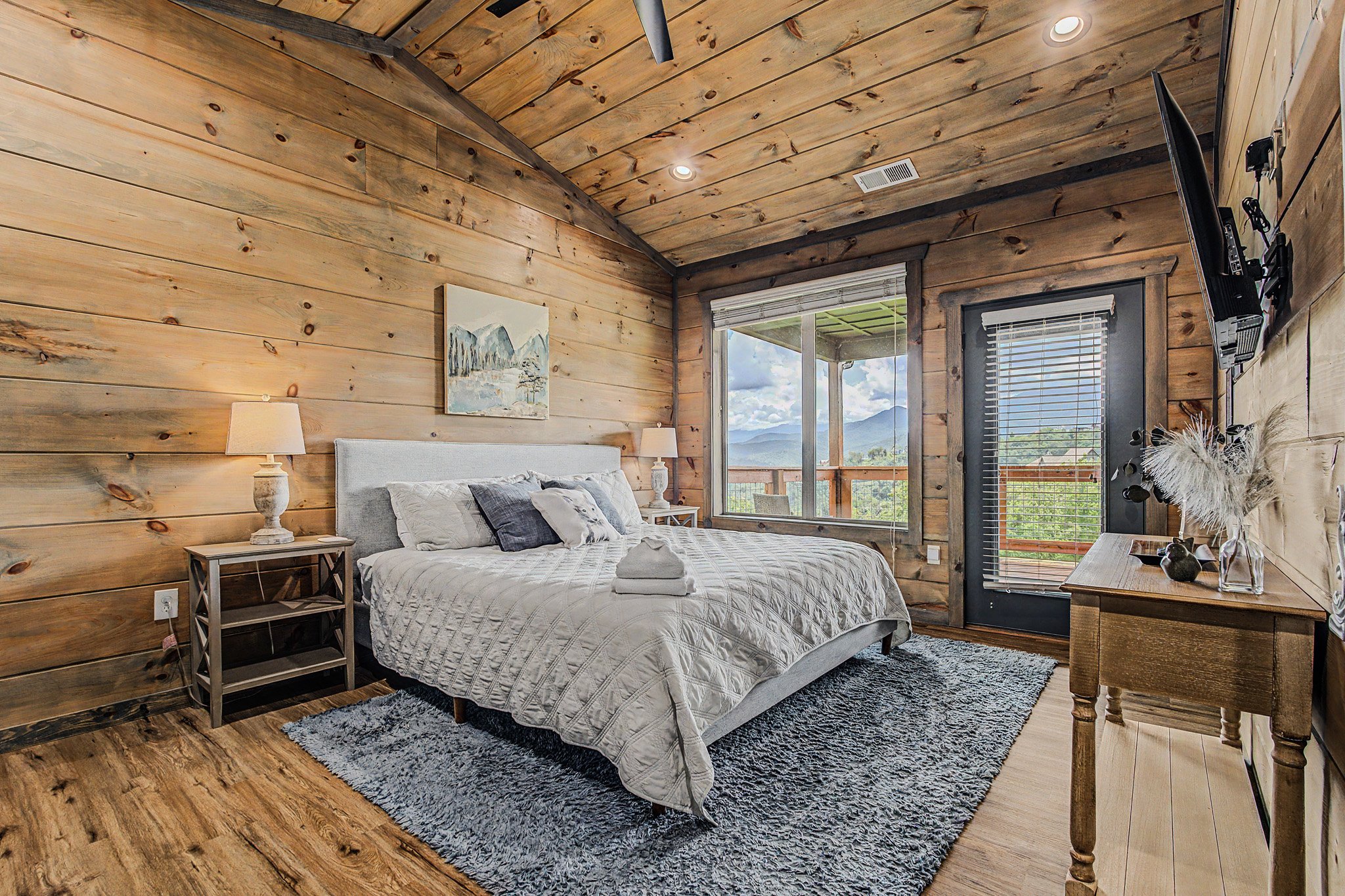 A rustic bedroom with wood-paneled walls and ceiling, a large window showing a mountain view, a bed with white bedding and decorative pillows, matching nightstands with lamps, and a wooden dresser with decorative items.