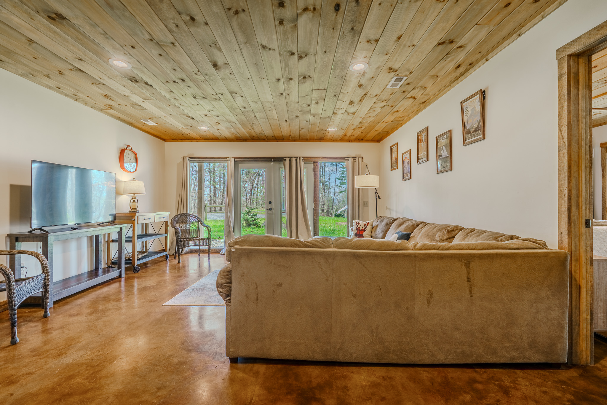 Living room with wooden ceiling, beige sofa, large glass doors opening to a green backyard, TV, and framed pictures on the wall.