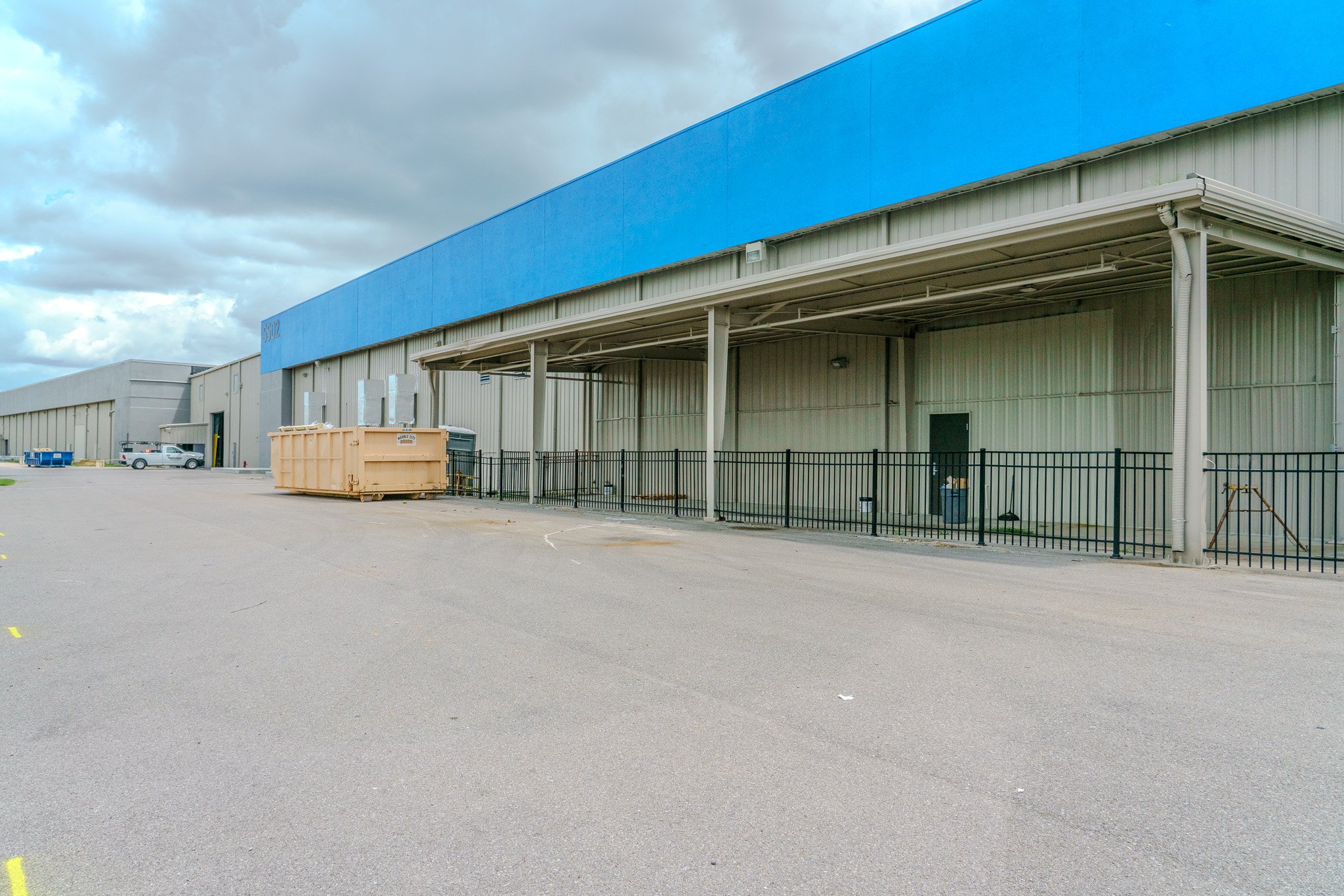 Exterior view of a large industrial warehouse with a blue stripe at the top, parking lot with few vehicles, and a large dumpster near the building.