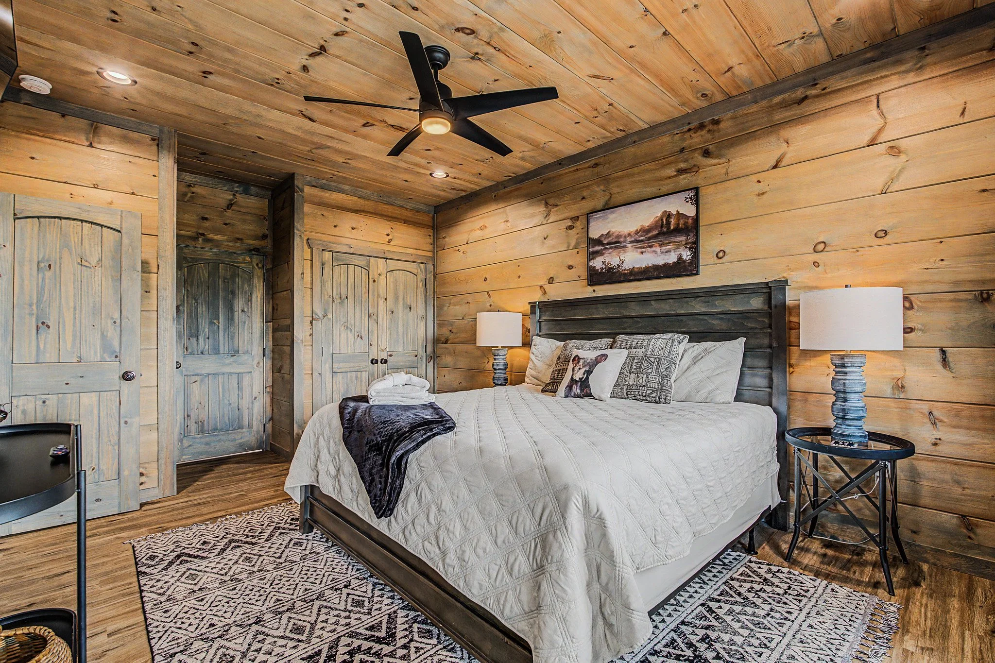 A bedroom with wooden paneled walls and ceiling, a bed with white bedding and decorative pillows, two matching white lamps on black side tables, a framed mountain landscape picture on the wall, and a black ceiling fan with a light in the center.