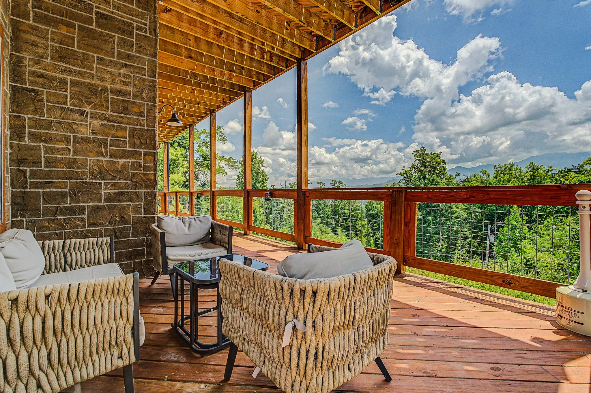 A balcony with wooden flooring, railing, and furniture including cushioned chairs and a glass table, overlooking a landscape of trees and mountains under a partly cloudy sky.