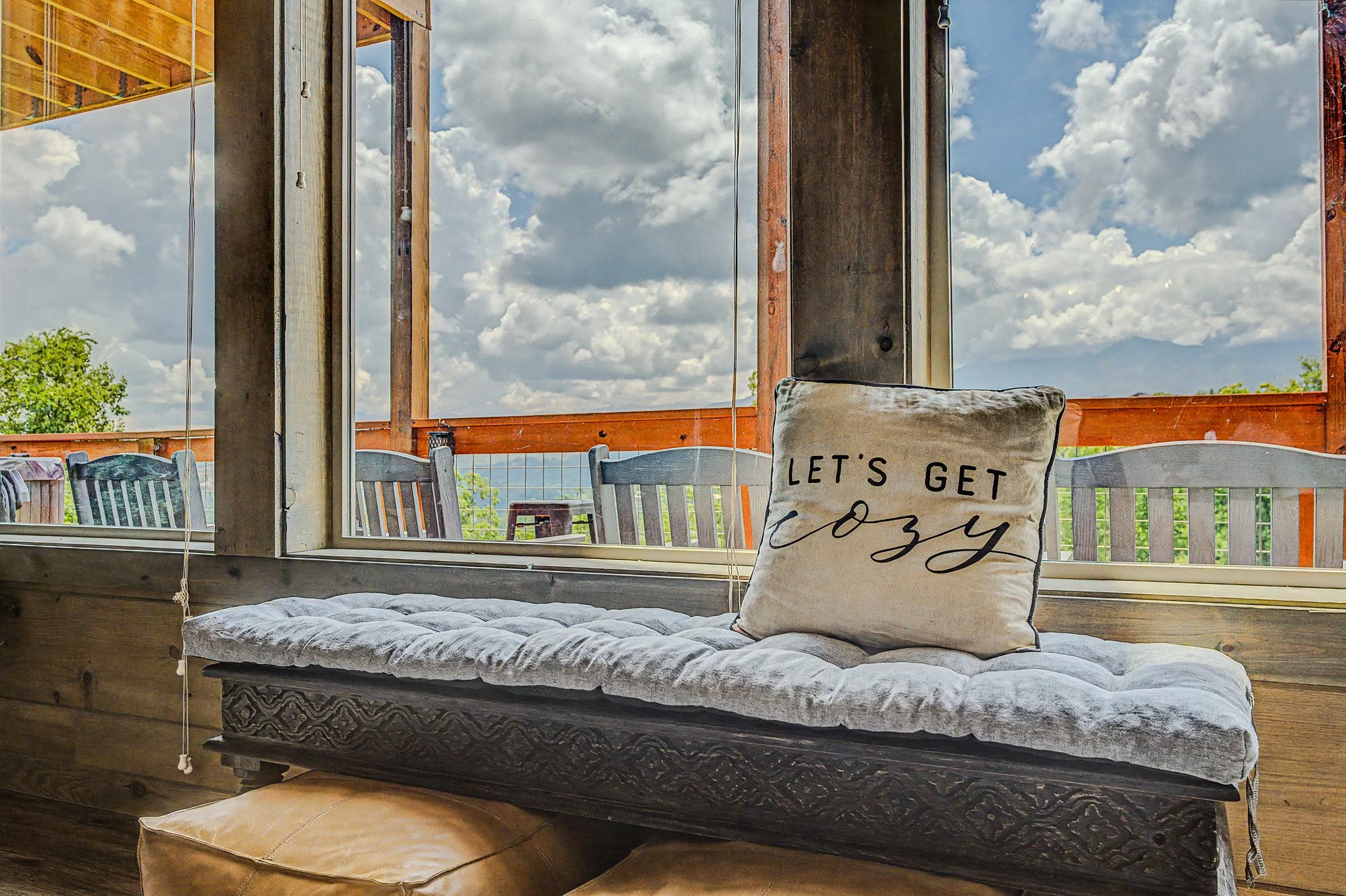 A cozy indoor seating area with a cushioned bench, a decorative throw pillow that reads 'Let's get cozy,' and large windows showing a partly cloudy sky and green trees outside.