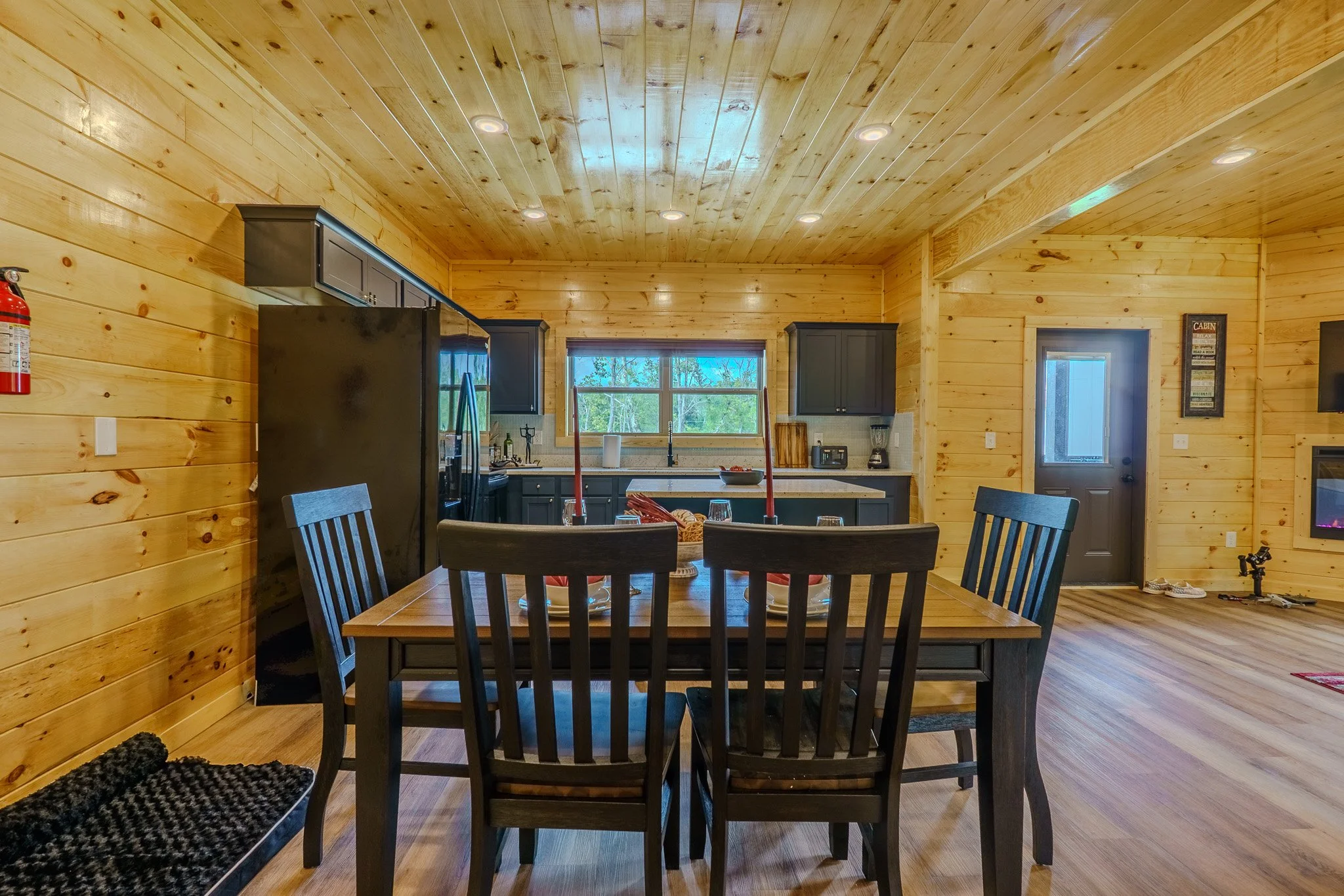 Interior of a wooden cabin with a dining table and chairs, kitchen area with black cabinetry, a window, and a door leading outside.