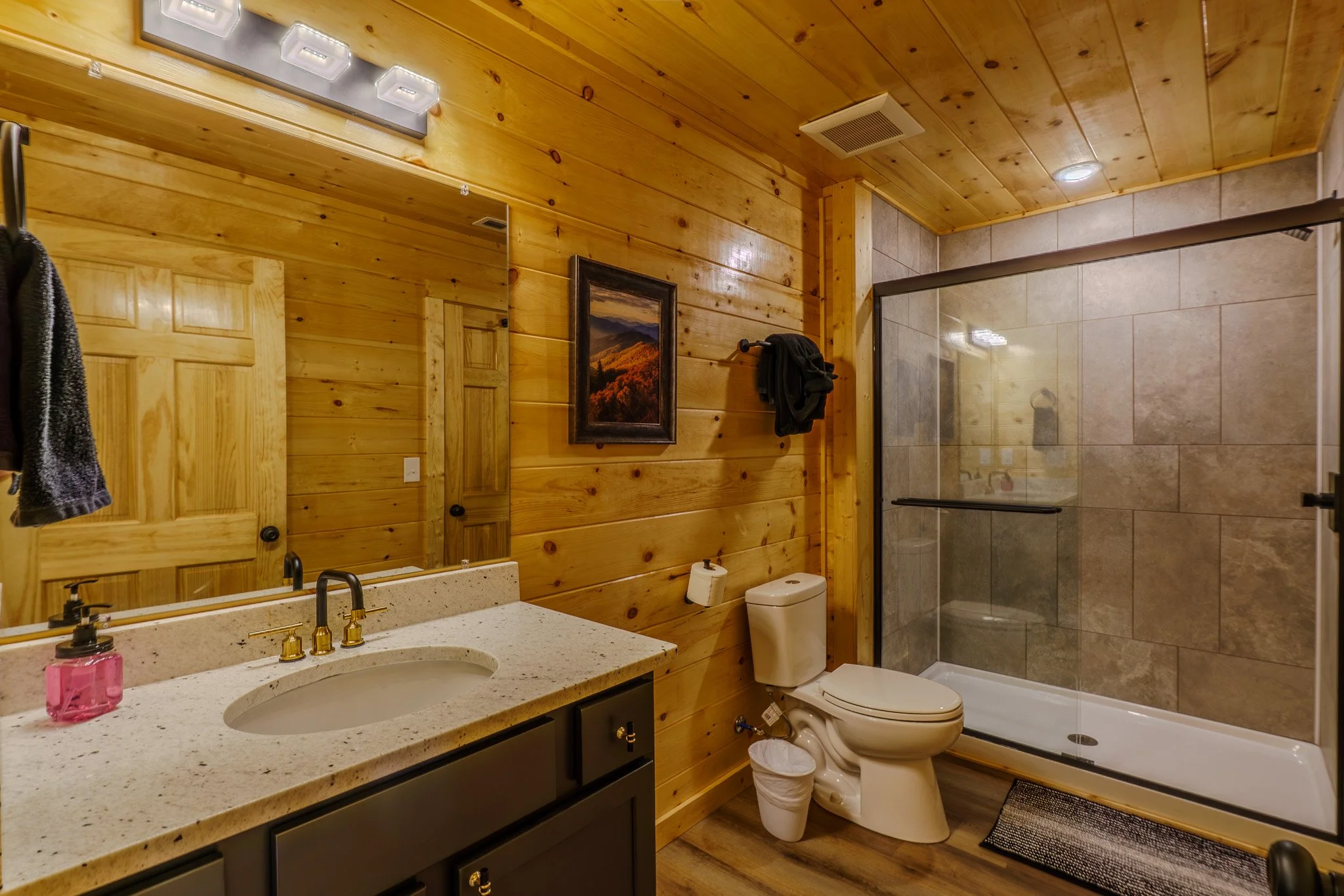 Bathroom with wood-paneled walls, a vanity with a sink and a mirror, a toilet, and a glass-enclosed shower with tiled walls.